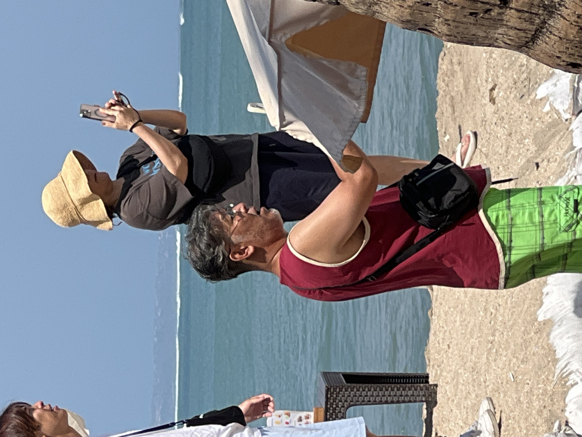 The image depicts a beach scene in Kuta, Indonesia, under a clear blue sky. In the foreground, a man with graying hair and glasses wears a red tank top and green plaid shorts, with a black fanny pack across his chest. He appears to be holding a light-colored beach umbrella. Behind him, a person wearing a wide-brimmed straw hat and a dark t-shirt is taking a photo with a smartphone. Another individual, partially visible on the left, wears a white shirt and light blue shorts. The background features the turquoise ocean with gentle waves and a distant coastline. A wicker-style side table is visible to the left of the man in red. The sand is coarse and light-colored.