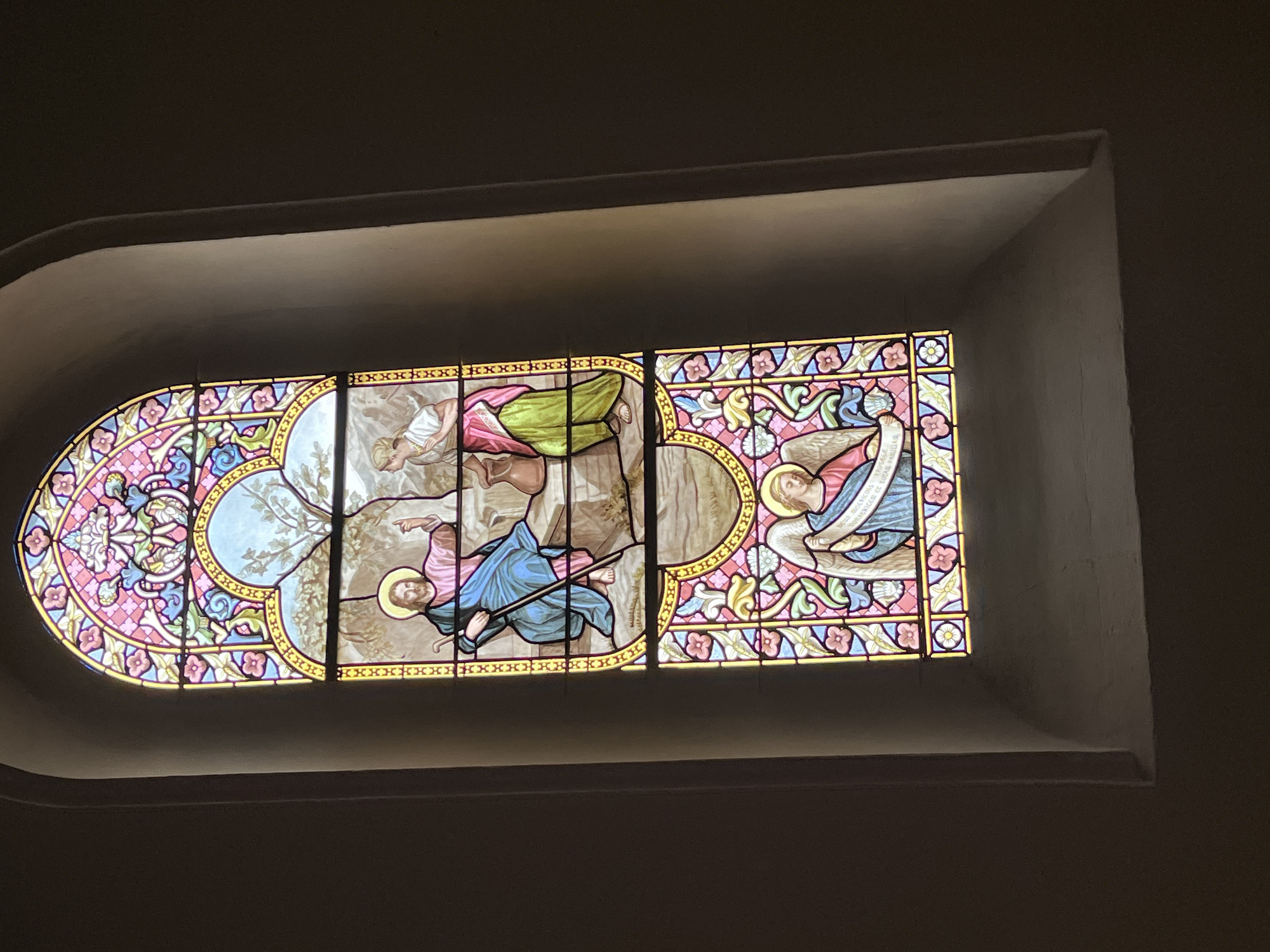 The image shows a stained glass window in what appears to be an indoor location, possibly a church, in Chambœuf, France. The window is comprised of three sections, each depicting biblical scenes with figures in colorful robes and haloes. The first section (left) features decorative floral designs along the sides. The second section displays two figures, one holding a staff. The third section (right) shows an angel. The background behind the stained glass is a dark wall, providing strong contrast. The window's frame is a light color.