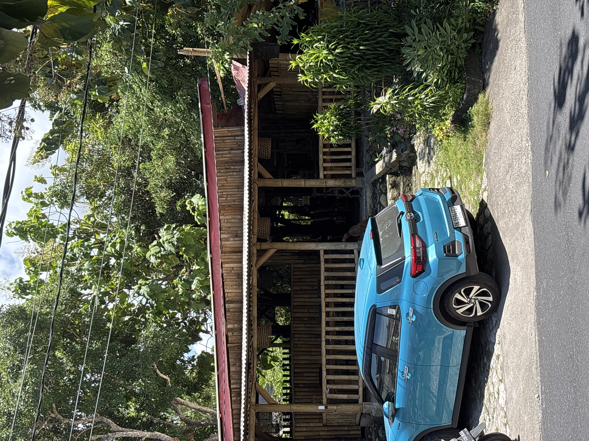 The image shows a blue Toyota Raize parked on the side of a narrow paved road next to a two-story bamboo structure. The vehicle has a license plate that reads "DK 1594 FQJ." The structure is open-sided with a red metal roof. Lush vegetation, including trees and bushes, surrounds the scene. The image was taken during daylight hours in Ungasan, Indonesia.
