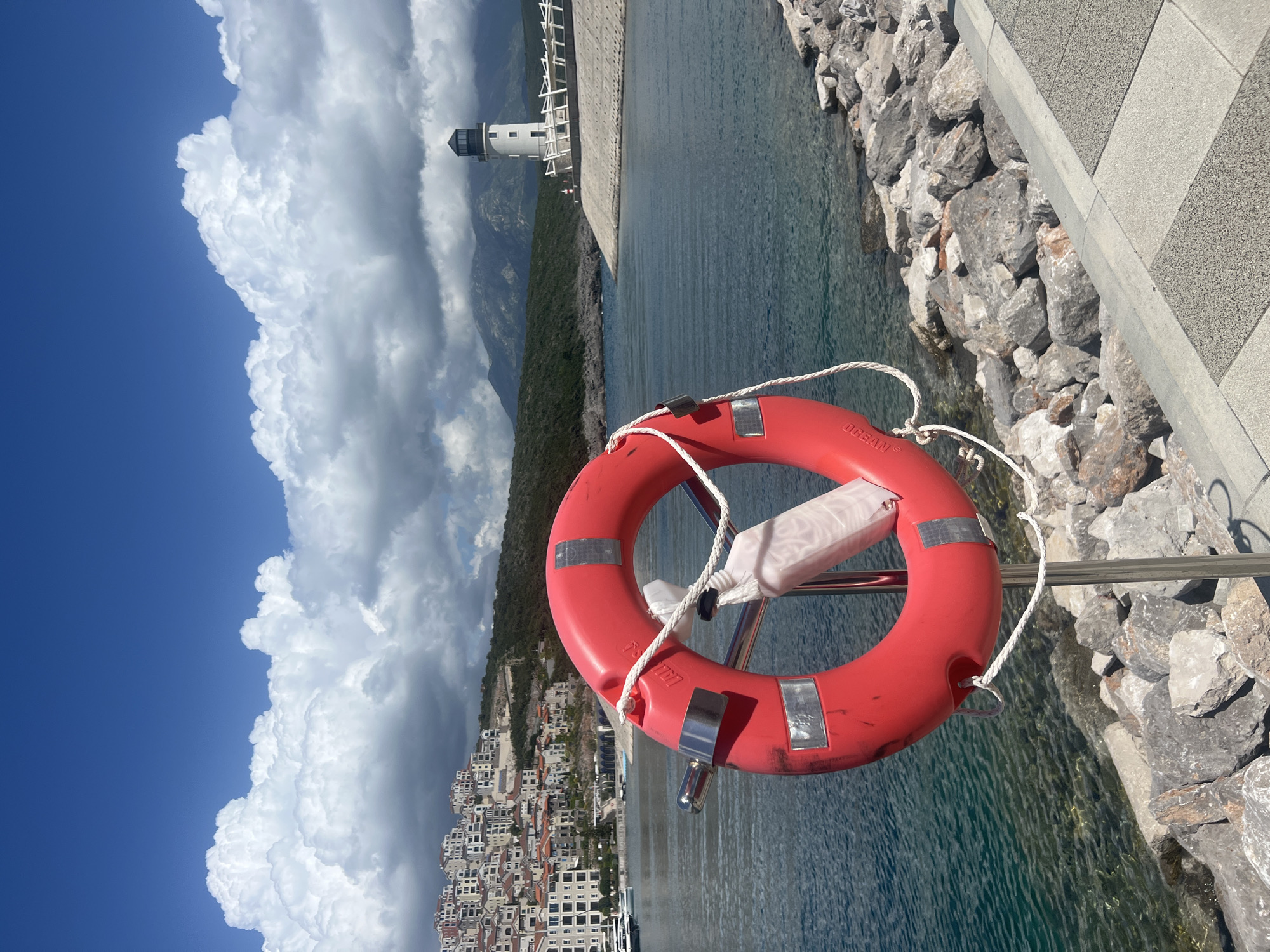 A medium shot shows a bright red life preserver with a white rope hanging from a chrome bar, against a landscape of water, rock, sky, and cityscape, in Radovići, Montenegro. The life preserver's labeling, "OCEAN," is partially visible. The water behind the life preserver is clear and turquoise, with visible underwater rocks near a jagged rock barrier. A gray paved walkway is adjacent to the rock barrier. In the distance, across the water, is a white lighthouse and a mountain range, with dense green vegetation. A dense cluster of buildings with red-tiled roofs is to the left of the mountains. The sky is partly cloudy, with a deep blue patch at the upper left, transitioning to large, fluffy white clouds.