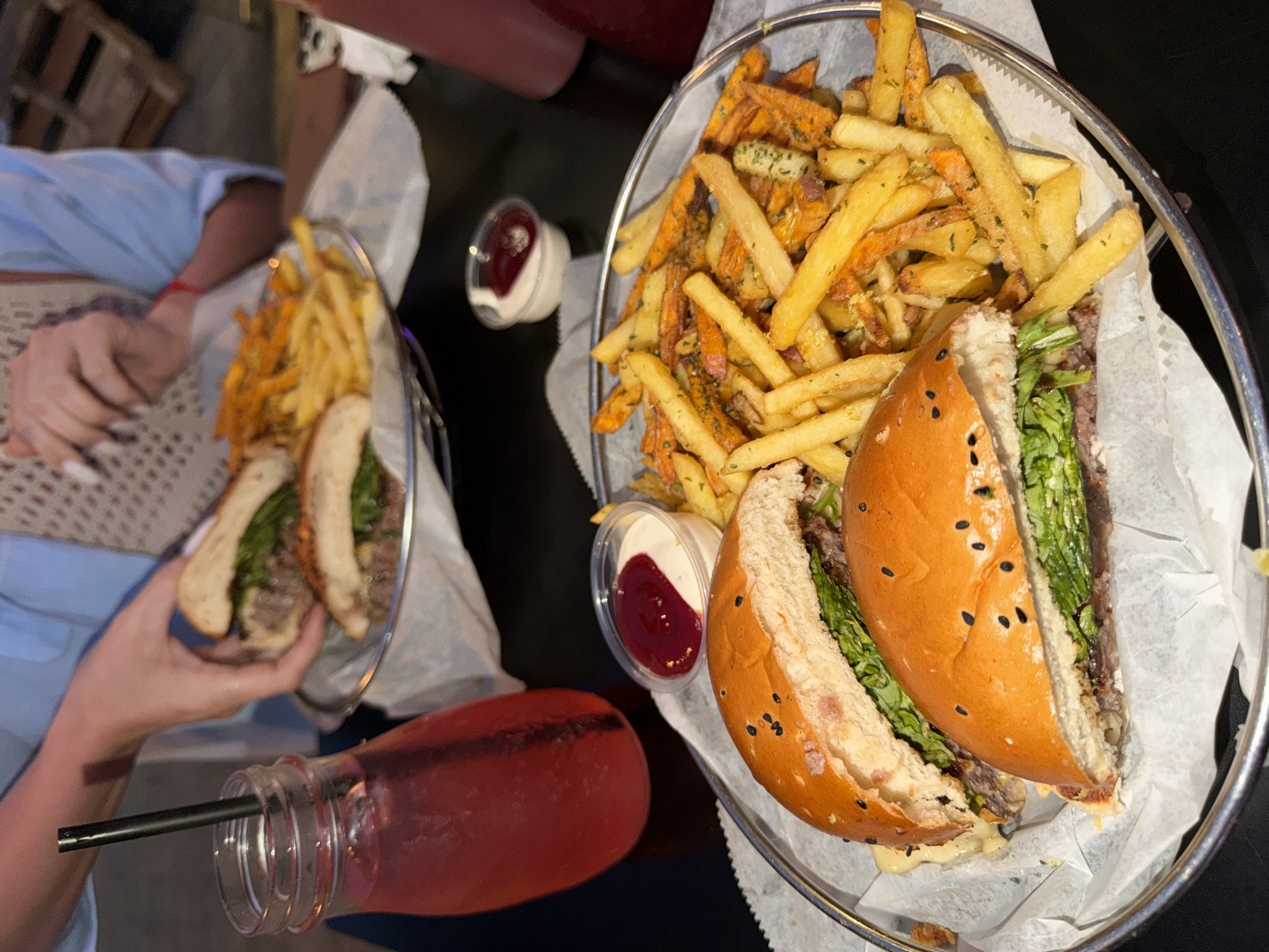 A top-down shot shows two meals each comprised of a cut burger and a mix of sweet potato and regular fries served in a metal basket lined with white paper. A small container with two-tone dressing sits beside each meal. In the lower left of the frame, a mason jar filled with red liquid is visible, including a black straw. A person on the left is holding a meal in one hand and has the other visible with long nails. The scene appears to be in a dimly lit restaurant. The location cannot be determined from the image.