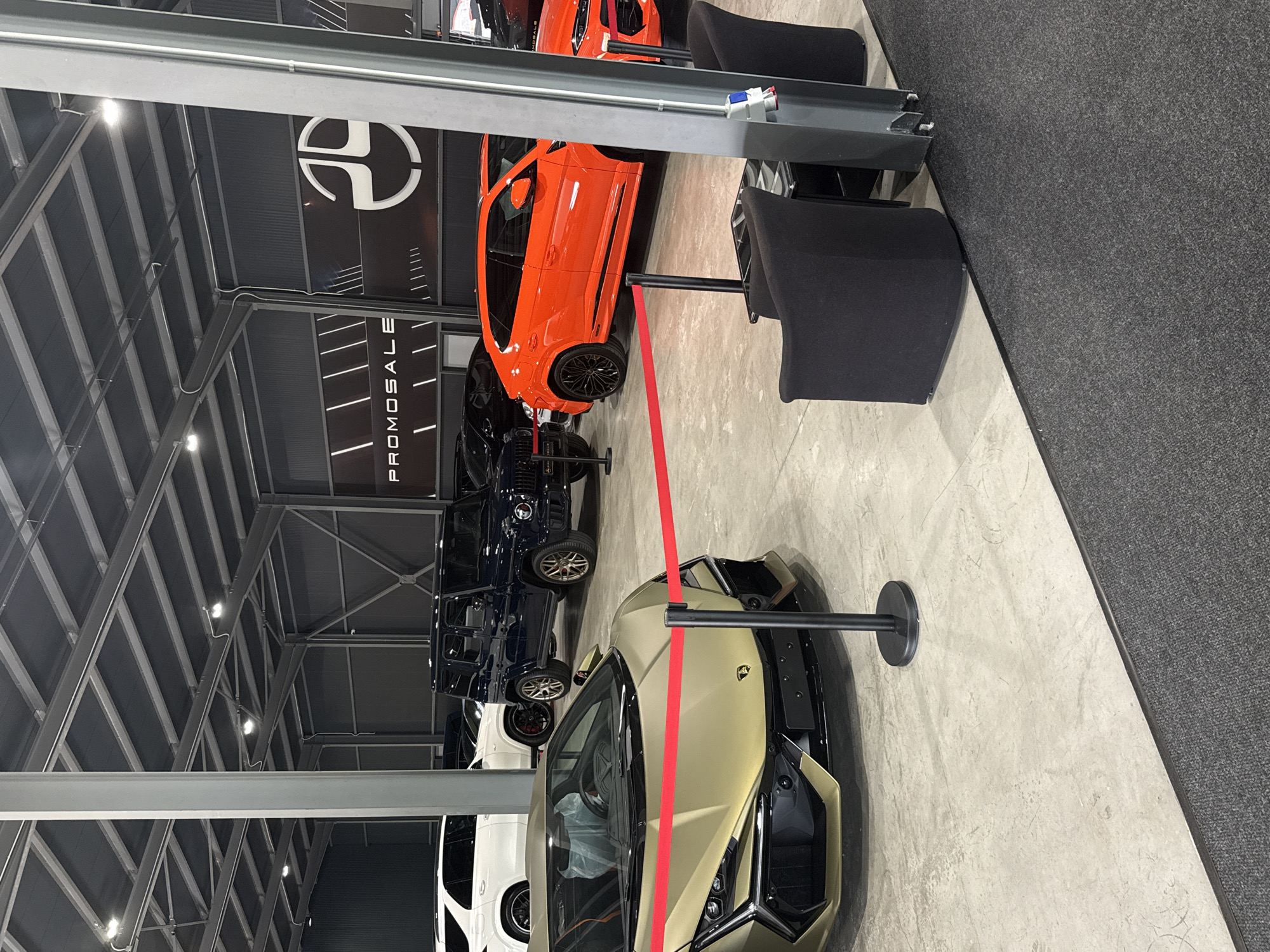 The image shows an indoor car showroom in Plovdiv, Bulgaria, featuring several luxury vehicles. The space is well-lit with a high, exposed metal framework ceiling. A prominent logo is visible on the wall, along with the word "PROMOSALE" vertically aligned. The vehicles are lined up, starting with a golden-colored Lamborghini in the foreground, followed by several black and dark blue SUVs, an orange sports car, and a white SUV in the background. A red barrier separates the display area from the pedestrian space. Two black chairs are situated near the orange car. The floor is concrete, with a gray carpet area to the right. The scene is static, without any people visible in the frame.