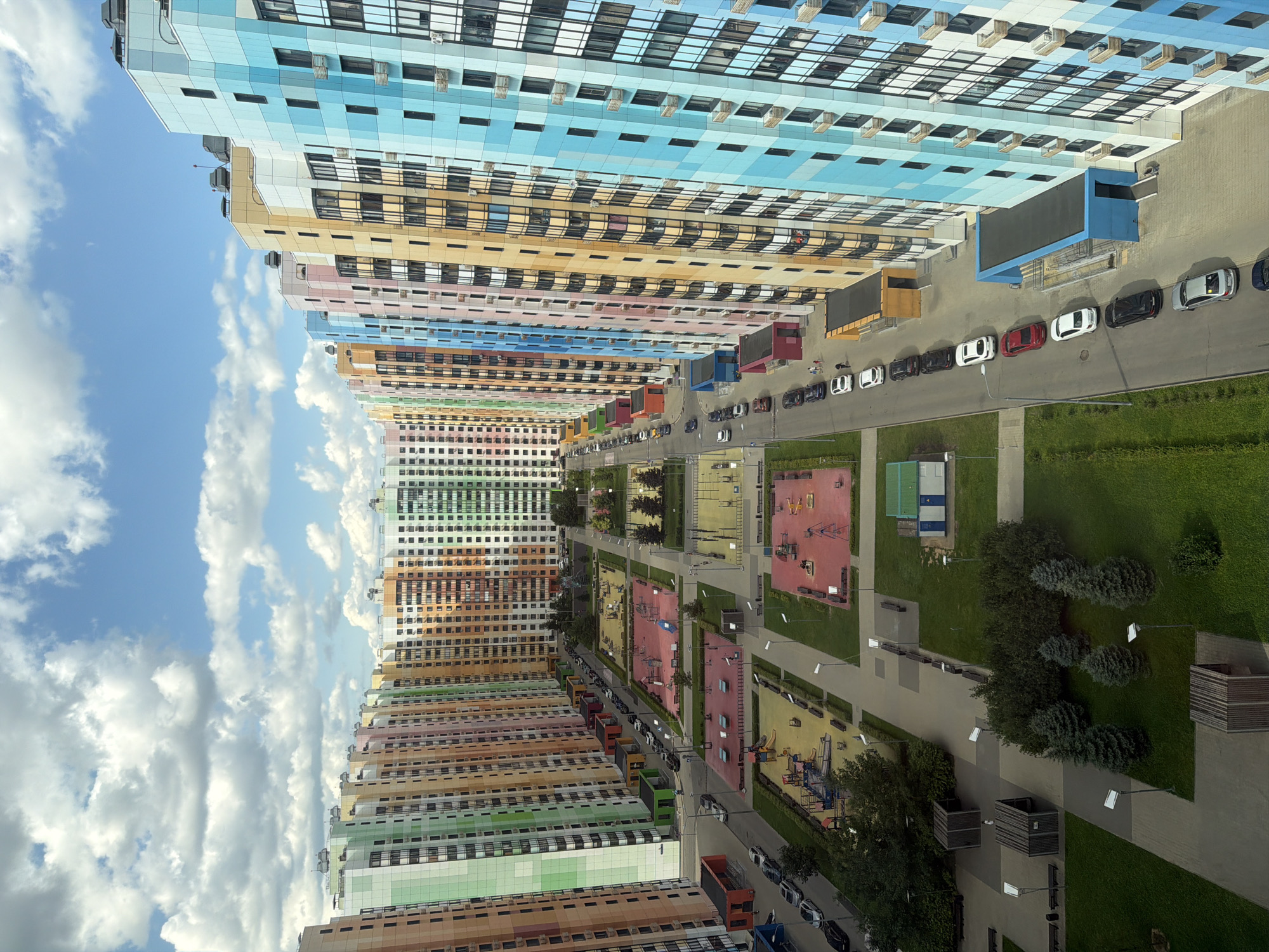 This is a high-angle, aerial shot of a modern, multi-colored residential complex under a partly cloudy sky. The buildings are tall, slender apartment blocks, painted in a vibrant array of colors, including pink, orange, yellow, green, blue, and white, arranged in a repeating pattern. The buildings line a central courtyard or open space. Within this open space are multiple playgrounds with red and yellow surfaces, featuring swings, slides, and climbing structures. There are also paved walkways, grassy areas, and several trees, including some small evergreen trees. A road runs along one side of the complex, lined with parked cars. The cars are of various colors, including black, white, silver, and red. A few people are visible walking on the sidewalks and near the playgrounds. The architecture suggests a contemporary urban development.