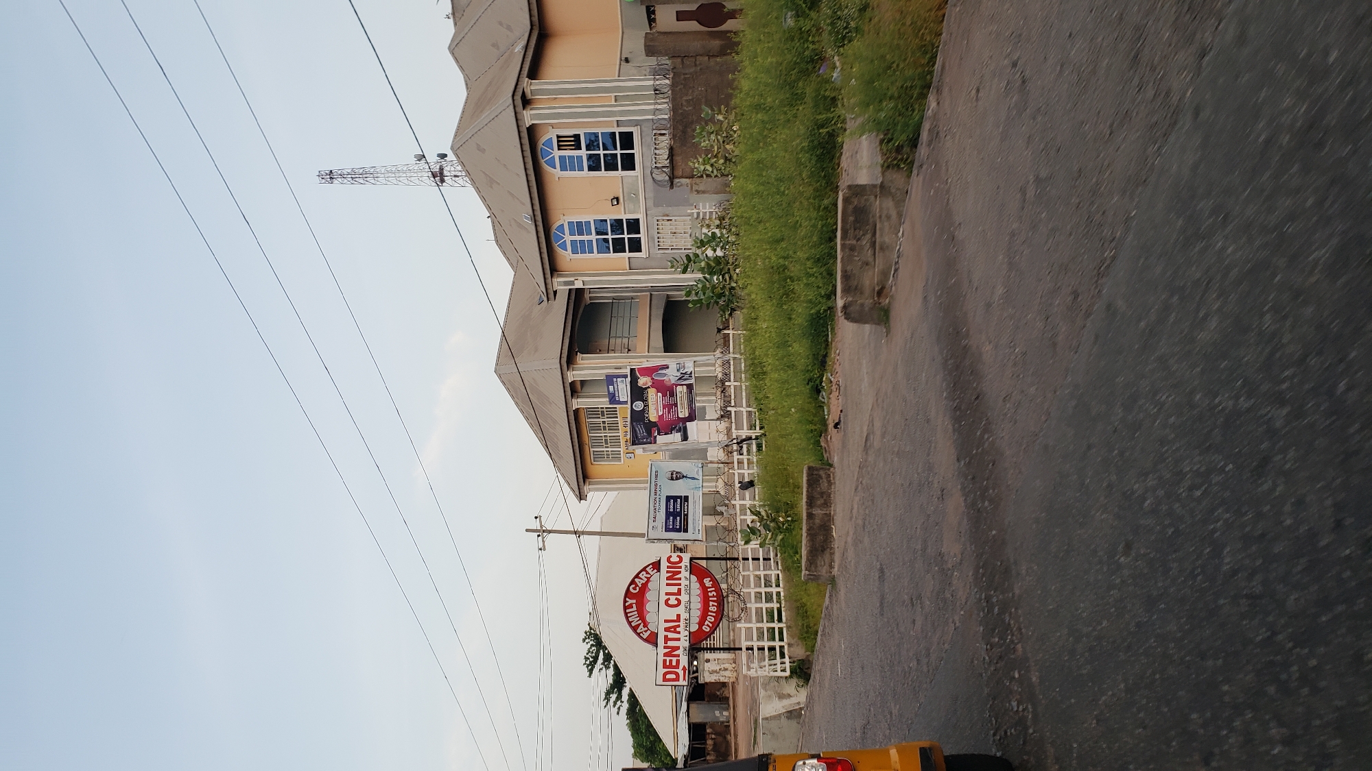 The image shows a street scene in Jalingo, Nigeria, featuring a dental clinic and residential buildings. In the foreground, a paved road shows signs of wear and tear, with some potholes and uneven surfaces. A yellow three-wheeled vehicle is partially visible on the left side of the frame.
Dominating the mid-ground is a red and white sign for "FAMILY CARE DENTAL CLINIC" with a phone number "07018715149". Behind the clinic sign, a white building with a sloped roof is visible. Adjacent to this, to the right, are two larger buildings. The first is a lighter-colored building with a balcony and several windows. The second building, to its right, is a two-story structure painted in yellow and cream colors, with arched windows on the upper floor. Several billboards are attached to the facade of these buildings, including one advertising "SALVATION MINISTRIES" with service times and another with images of people, possibly related to a church or ministry.
A white metal fence runs along the edge of the properties, and overgrown green grass and bushes are present in the landscape. In the background, a tall telecommunications tower stands against a pale blue sky with thin, horizontal power lines crossing the frame. The overall impression is of an urban environment in a developing country, with a mix of commercial and residential structures.