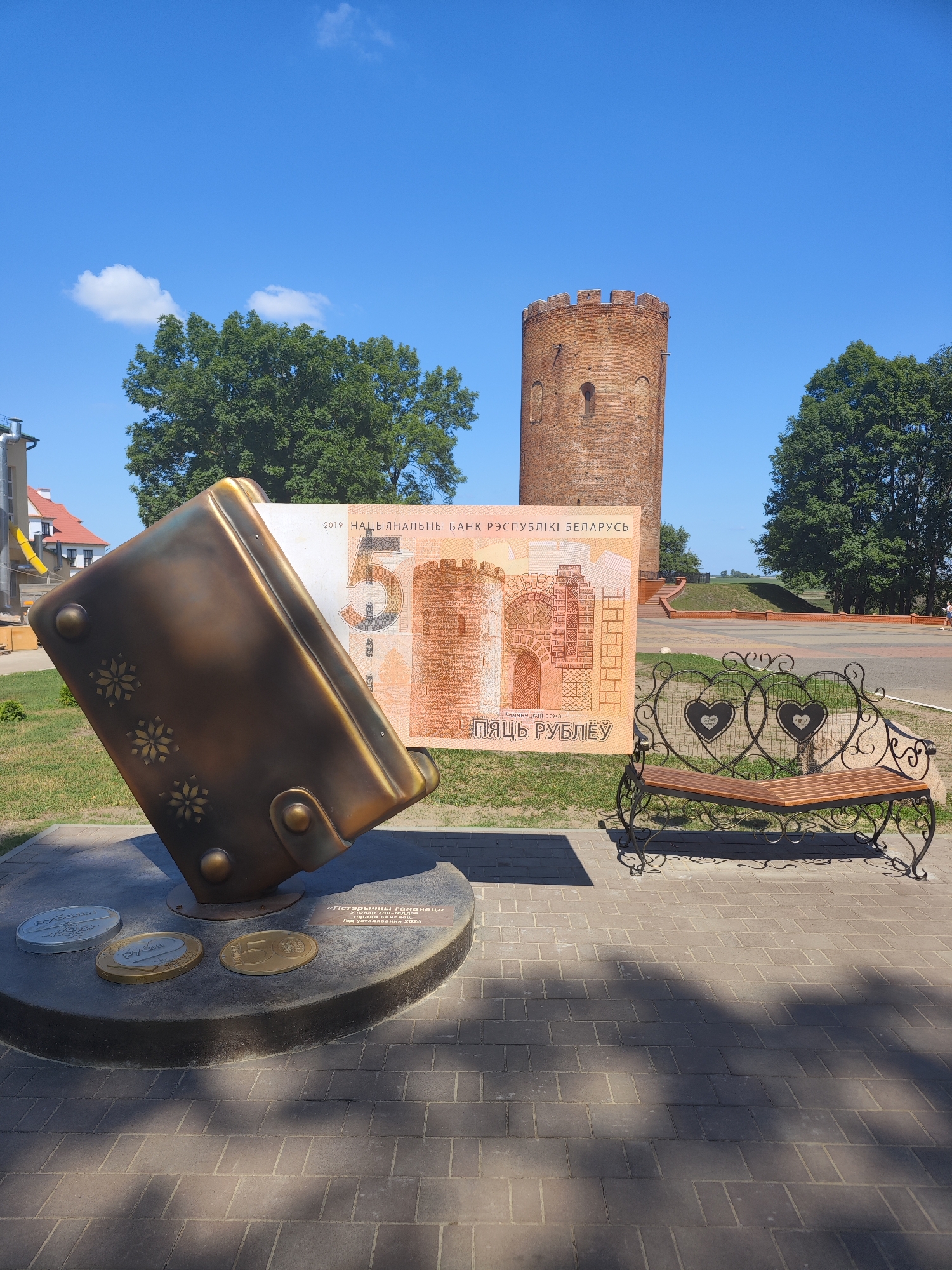This is a full outdoor, eye-level shot of a public space in Kamyanyets, Belarus on a sunny day with a clear blue sky. In the foreground, to the left, is a large bronze sculpture shaped like an open book, with a Belarusian 5 Ruble banknote placed inside it. The banknote features a depiction of the Kamyanyets Tower. Around the base of the sculpture are three oversized, coin-shaped elements, two with Russian text, and one with an image of a coin. To the right of the sculpture is an ornate black metal bench with a wooden seat, featuring heart designs. In the background, a historic brick tower, likely the Kamyanyets Tower itself, stands prominently against the blue sky. Lush green trees are visible behind the tower and to the right of the scene. The ground is paved with grey interlocking bricks, with patches of green grass surrounding the paved areas. There are no people clearly visible in the immediate vicinity of the main subjects.