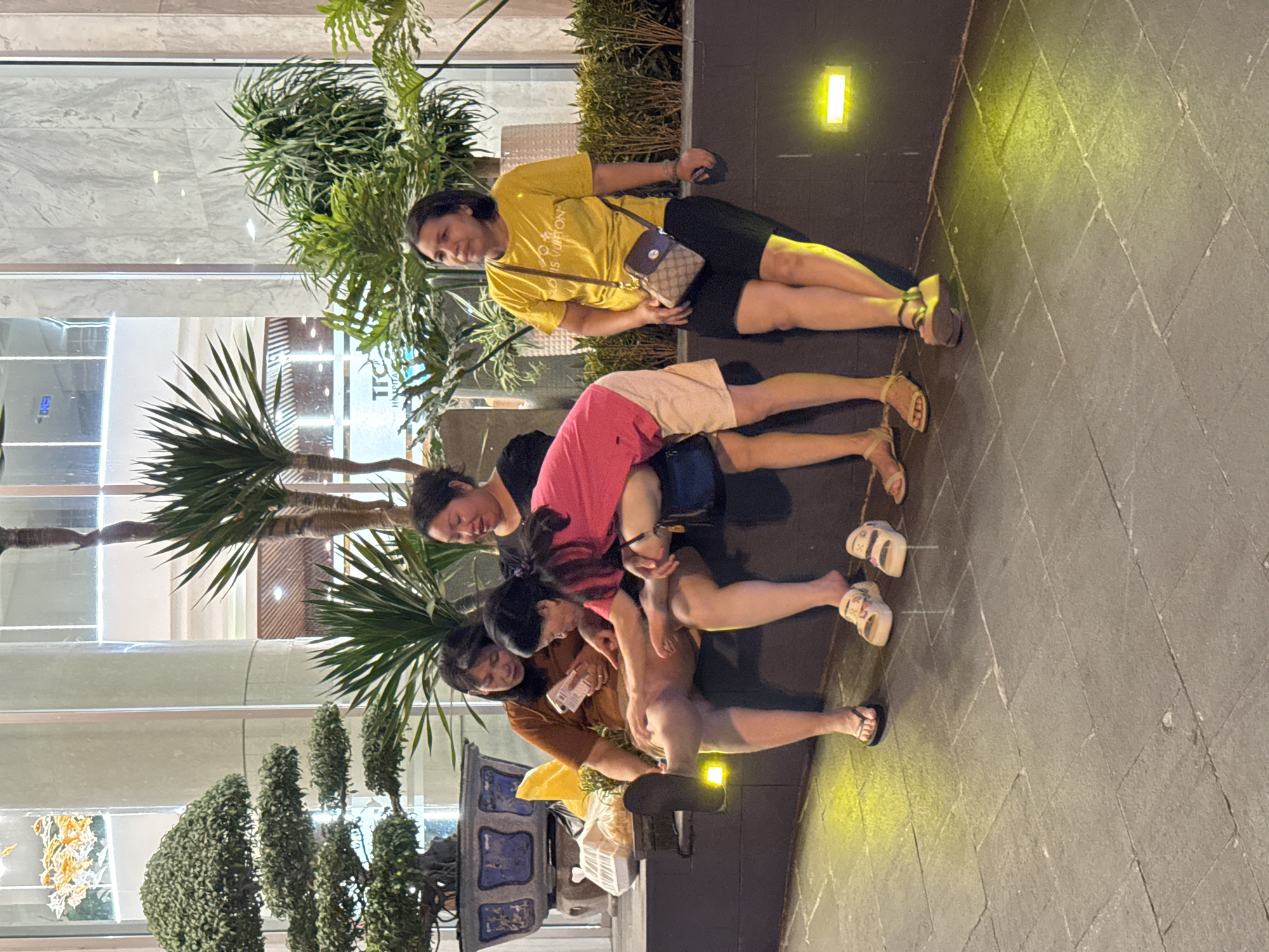 Four women are sitting on a low, dark-colored ledge in front of a building with large glass windows. The woman on the far left wears a brown t-shirt and tan shorts, with her left leg extended and wearing a black flip-flop. She is looking down at her phone. To her right, another woman in a pink t-shirt and white shorts is leaning over, looking at something on the lap of the woman next to her. The woman next to her, wearing a brown t-shirt, is looking down at her phone, and appears to be showing something to the woman in pink. The woman on the far right wears a yellow t-shirt and black shorts. She has a small brown and tan purse slung across her body and is looking off to her left. Behind the women, there are several large plants and trees. The ground in front of the ledge is paved with square tiles. Two yellow lights are visible on the dark wall behind the women.