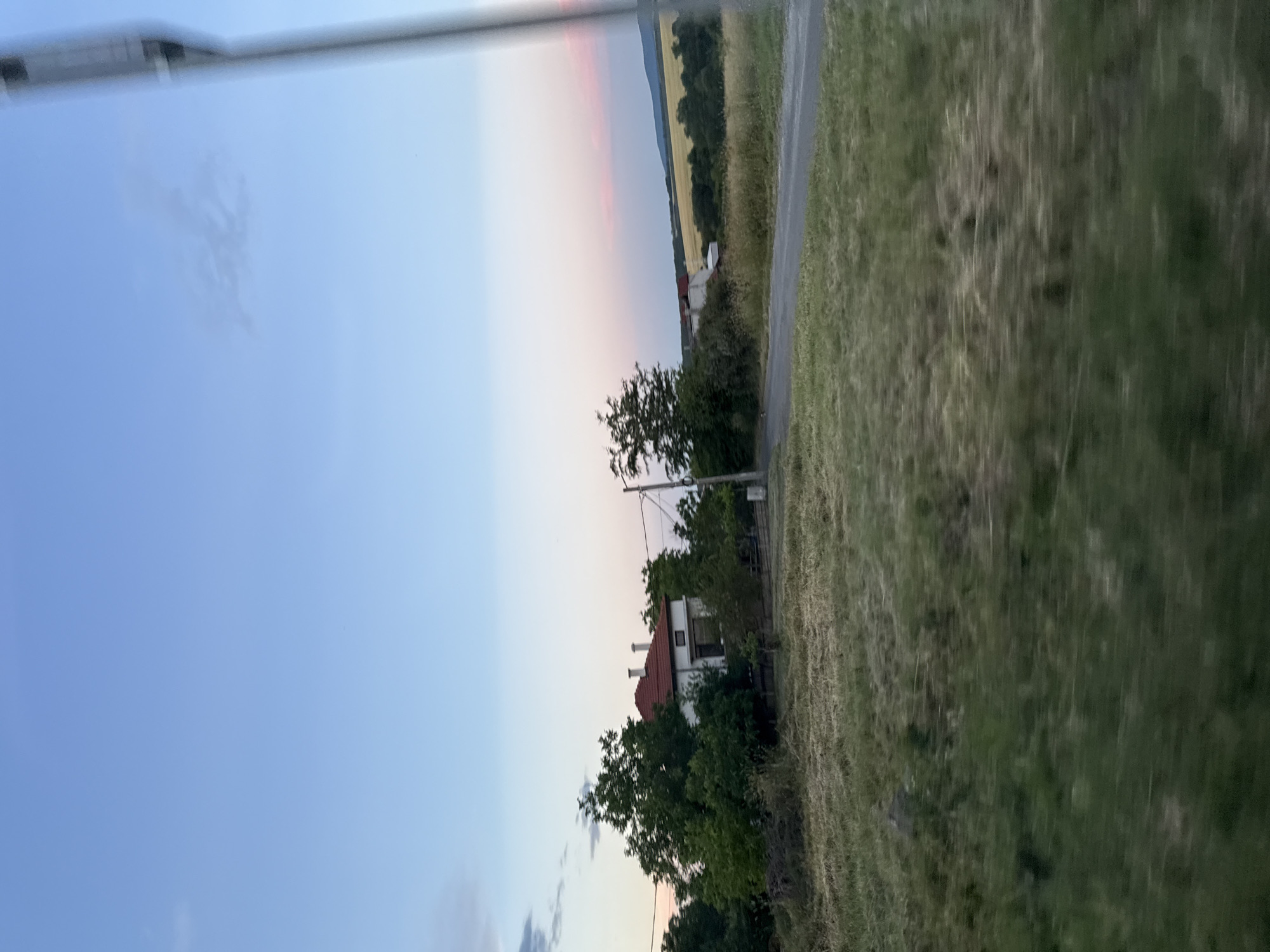 The image captures a rural scene with a house nestled amongst trees. The sky exhibits a gradient of blue fading into soft pinks and oranges, suggesting either dawn or dusk. A paved road runs through the foreground, bordered by dry grass and greenery. In the distance, a yellow field stretches towards rolling hills. A solitary lamp post stands to the right of the frame. The overall impression is one of quietude and natural beauty.