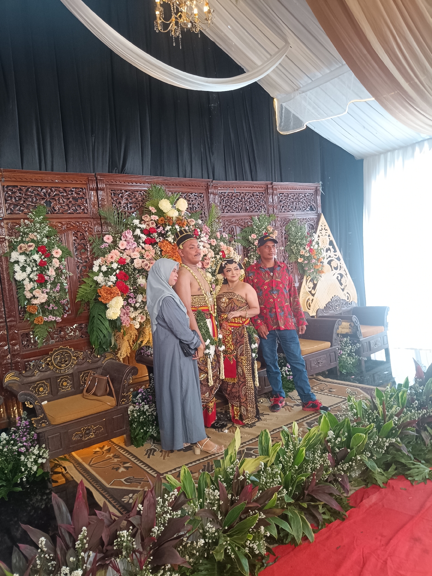 The image depicts a wedding ceremony staged indoors, likely at a venue in Depok, Indonesia. The setting is adorned with elaborate floral arrangements and traditional wooden backdrops. Four individuals are present: a bride and groom in traditional attire, a woman in a grey hijab and dress, and a man in a red patterned shirt and blue jeans. The bride and groom are positioned in the center, with the bride wearing a beaded headdress and a patterned dress, and the groom in a similar headdress and ceremonial garment. The woman in the hijab stands to the left, looking away from the camera, while the man in casual wear stands to the right. The scene appears to be a photo opportunity, with the subjects posing for the camera. The overall ambiance suggests a cultural and festive occasion.