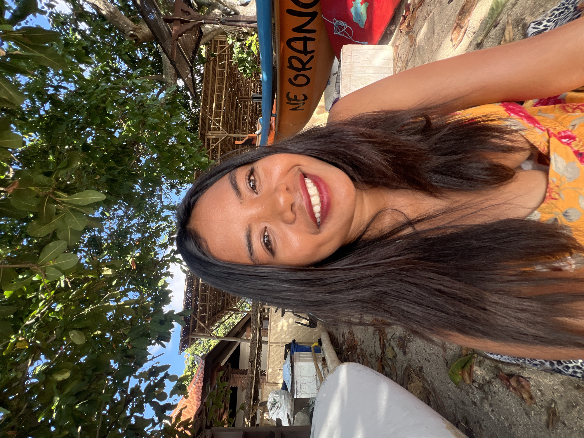 A portrait of a young woman with long, dark hair and fair skin, smiling at the camera. She is wearing a yellow and pink floral sundress. The background suggests a beachside location in Kuta, Indonesia. She is seated on the sand, with a white boat on her left and an orange boat with "NE ORANGE" written on it to her right. Behind her are trees, a thatched roof structure, and buildings. The overall atmosphere is casual and sunny.