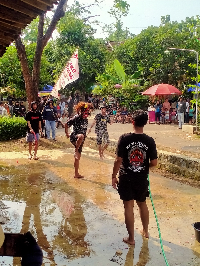 An outdoor cultural performance is captured in Gebyog, Indonesia, under clear daylight conditions. The scene unfolds on a wet, light-colored dirt or paved surface, reflecting the sky and surrounding elements. In the immediate foreground, a person with their back to the viewer wears a black t-shirt emblazoned with "SENI REOG TRADISIONAL JOKO REWANG" and black shorts, standing barefoot while holding a green garden hose that extends towards a black bucket on the far right.
Three central figures are actively engaged in the mid-ground, all barefoot and standing in shallow water. A performer on the left wears a black t-shirt, dark shorts, and a red, multi-colored mask or face paint, with visible brown dreadlocks. This individual is depicted in a dynamic pose, lifting their right knee high. To their right, another male performer, wearing a dark patterned t-shirt and dark shorts, appears to be splashing water, with visible water droplets around him. Further left, a third male figure in a black hoodie and light-colored shorts holds a red whip-like object.
In the background, a crowd of approximately 30-40 spectators, consisting of adults and children, observes the performance. Some individuals are standing, while others are seated directly on the ground. Green foliage from numerous trees dominates the background, with a distinct banana tree visible on the right. A large white flag with red text, partially legible as "JOKO ENAK," is attached to a pole on the left. On the right side of the background, a red market umbrella covers what appears to be a small stall or vendor area, with several people standing nearby. The overall environment suggests a festive or ceremonial gathering.