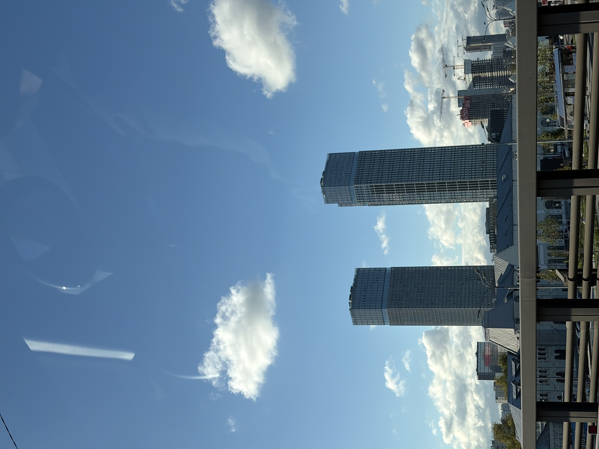 Two modern, identical skyscrapers with glass facades and dark grid patterns dominate the center of the frame, standing against a bright blue sky with scattered white cumulus clouds. In the foreground, a grey metal railing suggests the perspective is from a bridge or elevated walkway. Beyond the skyscrapers, a cluster of construction cranes and partially built structures are visible, indicating ongoing development in the area. Lower buildings with grey, pitched roofs and ornate architectural details are situated in the middle ground, appearing to be older architecture. The overall scene depicts a blend of modern and older urban architecture under a clear sky, likely in a developing district of Moscow, Russia.