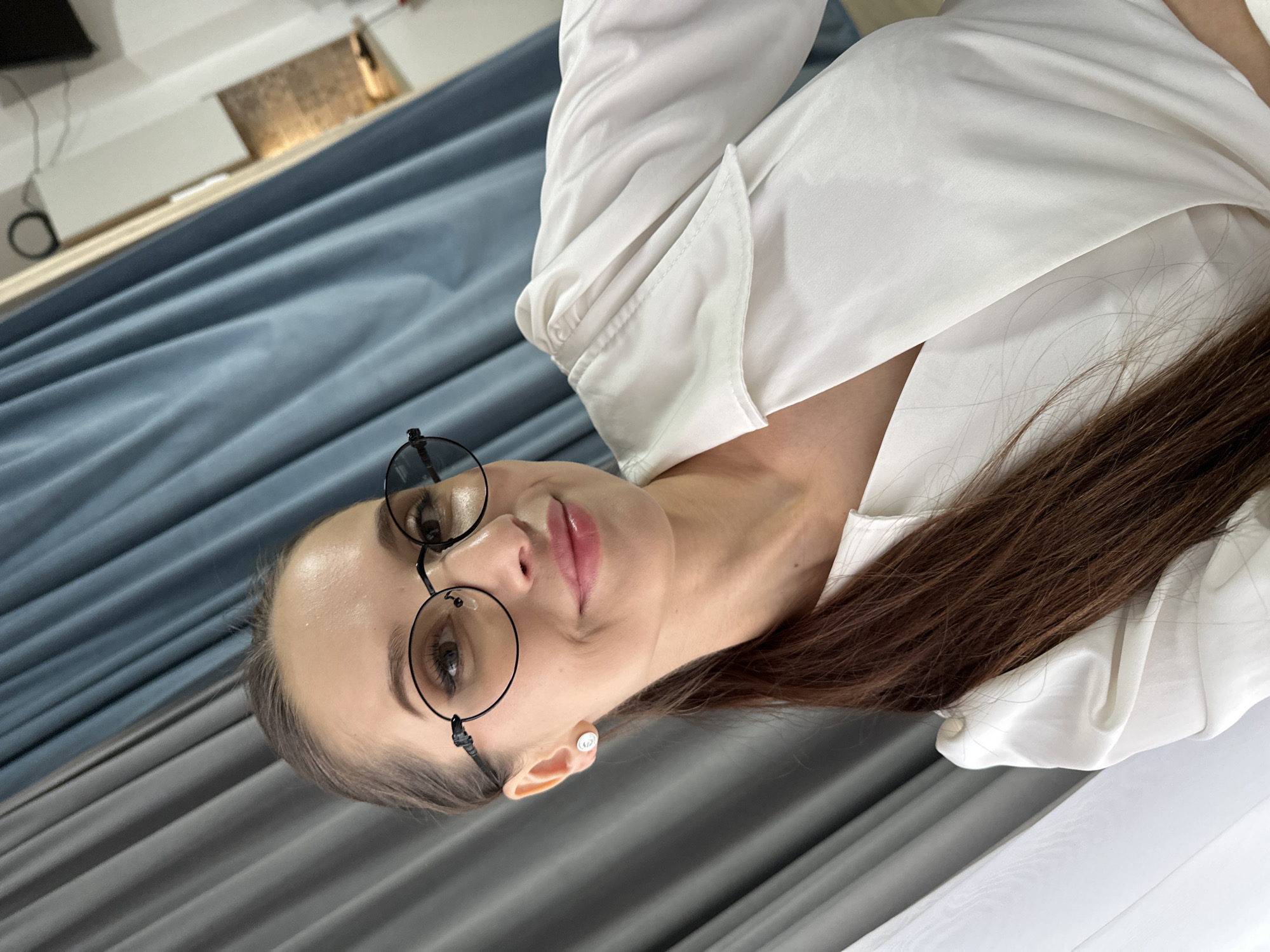 A portrait-style shot features a young woman looking directly at the camera. She has long, straight brown hair and is wearing a white collared shirt with visible sleeves. Her face is in clear focus, with well-defined eyebrows, open eyes, and slightly parted, pink-toned lips. She wears round, dark-rimmed glasses and a small, circular, silver-colored earring with an indistinct symbol in her right ear.
The background consists of draped fabric, likely curtains, with prominent blue sections on the left and grey sections on the right, creating a soft, textured backdrop. Beyond the curtains, a blurred indoor setting with some architectural or furniture elements is visible.
The woman's expression is calm and gentle, indicating a relaxed pose for the photograph. The lighting is soft and even, highlighting her features. The scene takes place indoors, potentially in a home or office, within Yekaterinburg, Russia. There is no discernible text visible in the image beyond the indistinct symbol on the earring.