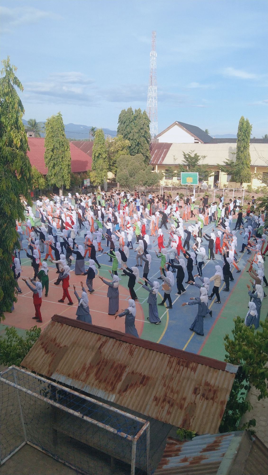 In Rhieng Blang, Indonesia, a large group of students, predominantly female and dressed in school uniforms including hijabs, are engaged in a synchronized physical exercise routine on an outdoor court. The court is marked with various colored lines, suggesting it may be a basketball or volleyball court. The students are spread across a significant portion of the court, performing arm movements in unison. Some students are in the foreground, clearly visible, while others are further back, appearing as a dense crowd. The surrounding environment includes school buildings with red-tiled roofs, lush green trees, and a tall telecommunications tower in the distance. The sky is partly cloudy, indicating daytime. In the foreground, a rusty corrugated metal roof and part of a soccer goal with netting are visible, suggesting the vantage point is from an elevated position overlooking the school grounds. The overall scene depicts a school activity, likely a morning exercise session.