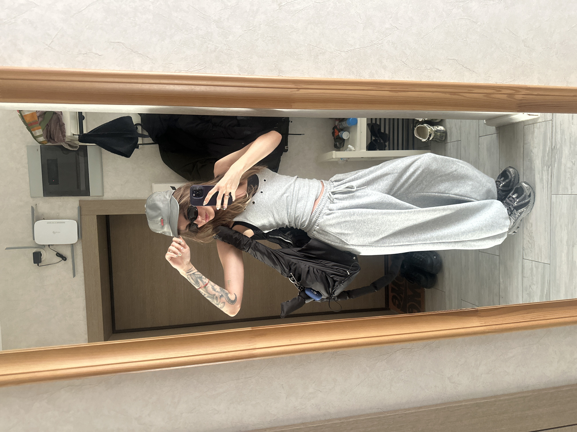 A young woman is taking a mirror selfie in an apartment entryway. She is wearing a gray baseball cap, sunglasses, a gray tank top, and baggy gray sweatpants. A black backpack is slung over her left shoulder. Her right arm is raised, and she is holding her phone to take the picture. Her left arm is bent at the elbow, and her hand rests on her hip. The background shows a wall with a router and shelf mounted on it, a coat rack with clothing, and a shoe rack with several pairs of shoes. The floor is tiled. The mirror is framed with light wood. The overall scene suggests a casual, everyday moment. The location is identified as an apartment in Moscow, Russia.
