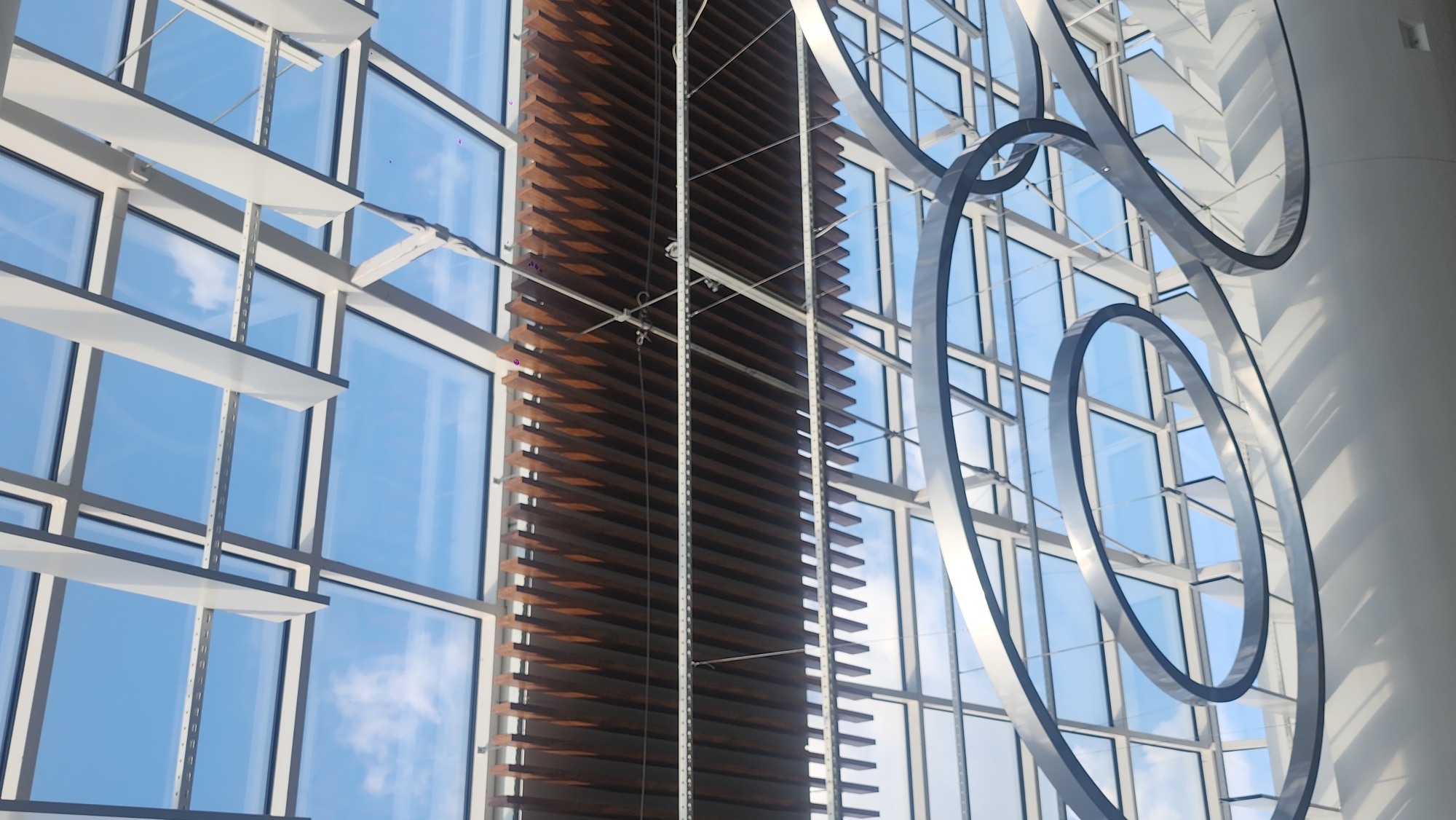 The image captures an interior view looking upwards towards a modern ceiling structure with a glass roof. The sky, visible through the glass panels, is a bright blue with scattered white clouds. The ceiling is comprised of a complex framework of white beams and panels. Below this, a wooden slatted ceiling element is suspended, casting a patterned shadow. Further down, two large, abstract, metallic ring sculptures are prominently featured, casting dramatic shadows on a white surface below. The overall impression is one of contemporary architecture and design.