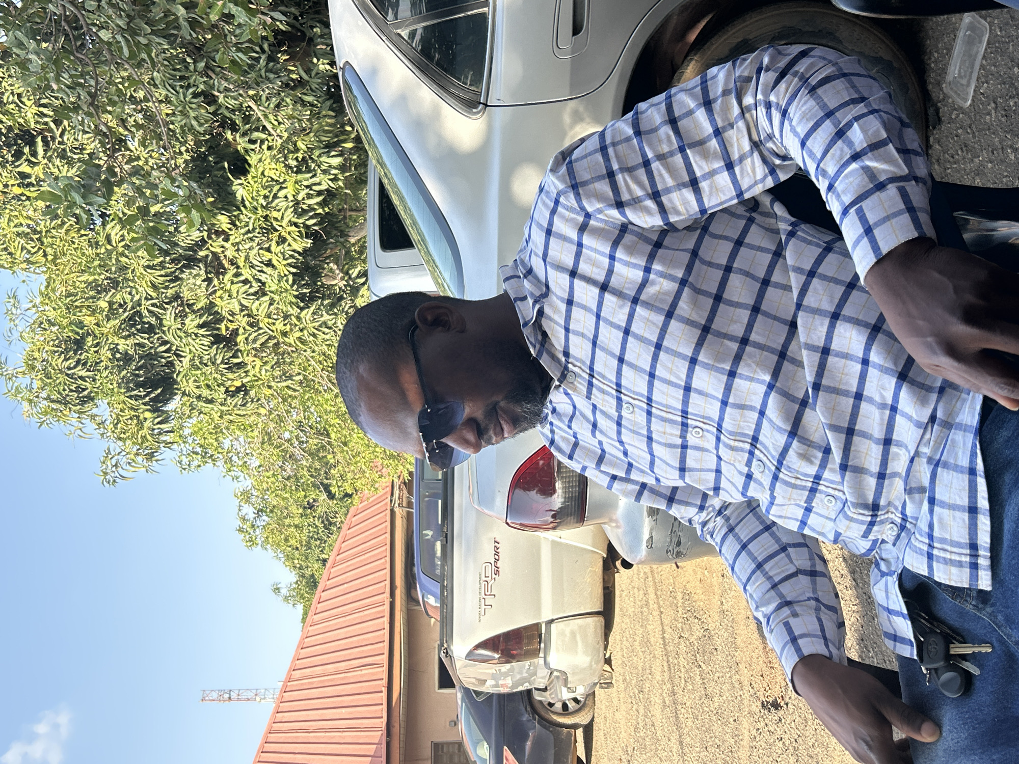 This image, captured outdoors in Jos, Nigeria, features an adult male seated in the foreground under clear daylight conditions. The man has dark skin, a short beard, and is wearing dark sunglasses, a blue and white plaid long-sleeved shirt, and blue jeans. A set of keys, including a Toyota car key fob, rests on his lap. He is looking slightly to his right and downward. The background reveals an unpaved ground area where several vehicles are parked. A prominent silver Toyota Tacoma pickup truck, displaying "TOYOTA RACING DEVELOPMENT TRD SPORT" on its tailgate, is centrally located. To its right, the rear section of a silver sedan is visible, and to its left, parts of a dark-colored sedan and a blue vehicle are seen. Behind the vehicles, a building with a reddish-brown corrugated metal roof stands. Dense green foliage from multiple trees dominates the right side and upper background. In the far left, a tall metal lattice tower is silhouetted against a clear blue sky, which features one small white cloud. The scene suggests a casual, semi-urban or rural-fringe environment.