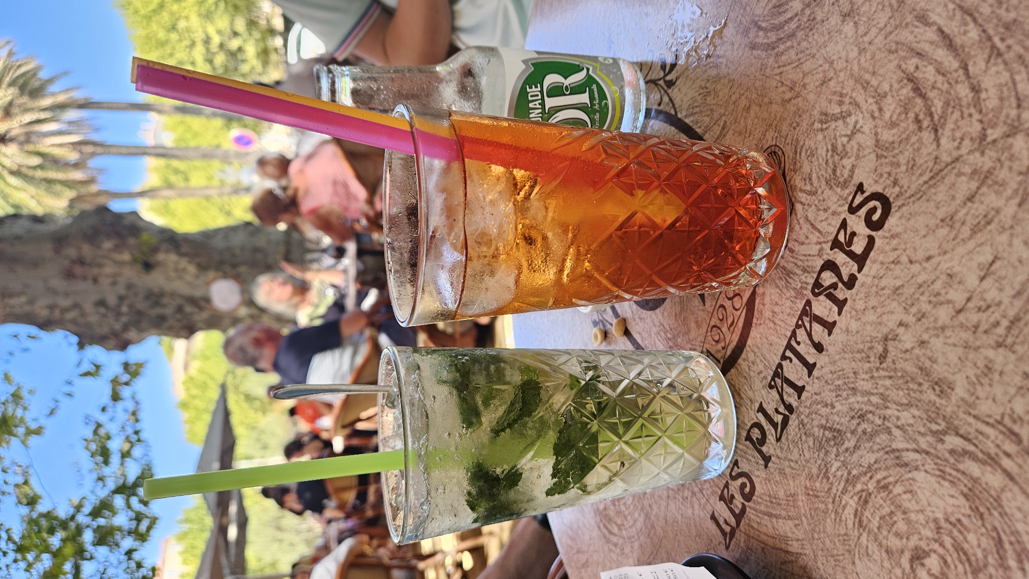 Two drinks are on a patterned table. The drink on the left is a clear cocktail with mint leaves and ice, with a green straw. The drink on the right is a reddish-orange cocktail with ice and orange garnish, with pink and yellow straws. A bottle of sparkling lemonade is behind the right drink. In the background, people are seated at tables under umbrellas, with trees and a blue sky visible. The table has the words "LES PLATANES 1928" printed on it. This is likely a cafe or restaurant in L'Île-Rousse, Corsica, France, as "Les Platanes" is a common name for establishments in the region, and the presence of palm trees and the relaxed outdoor setting are characteristic of Mediterranean coastal towns.