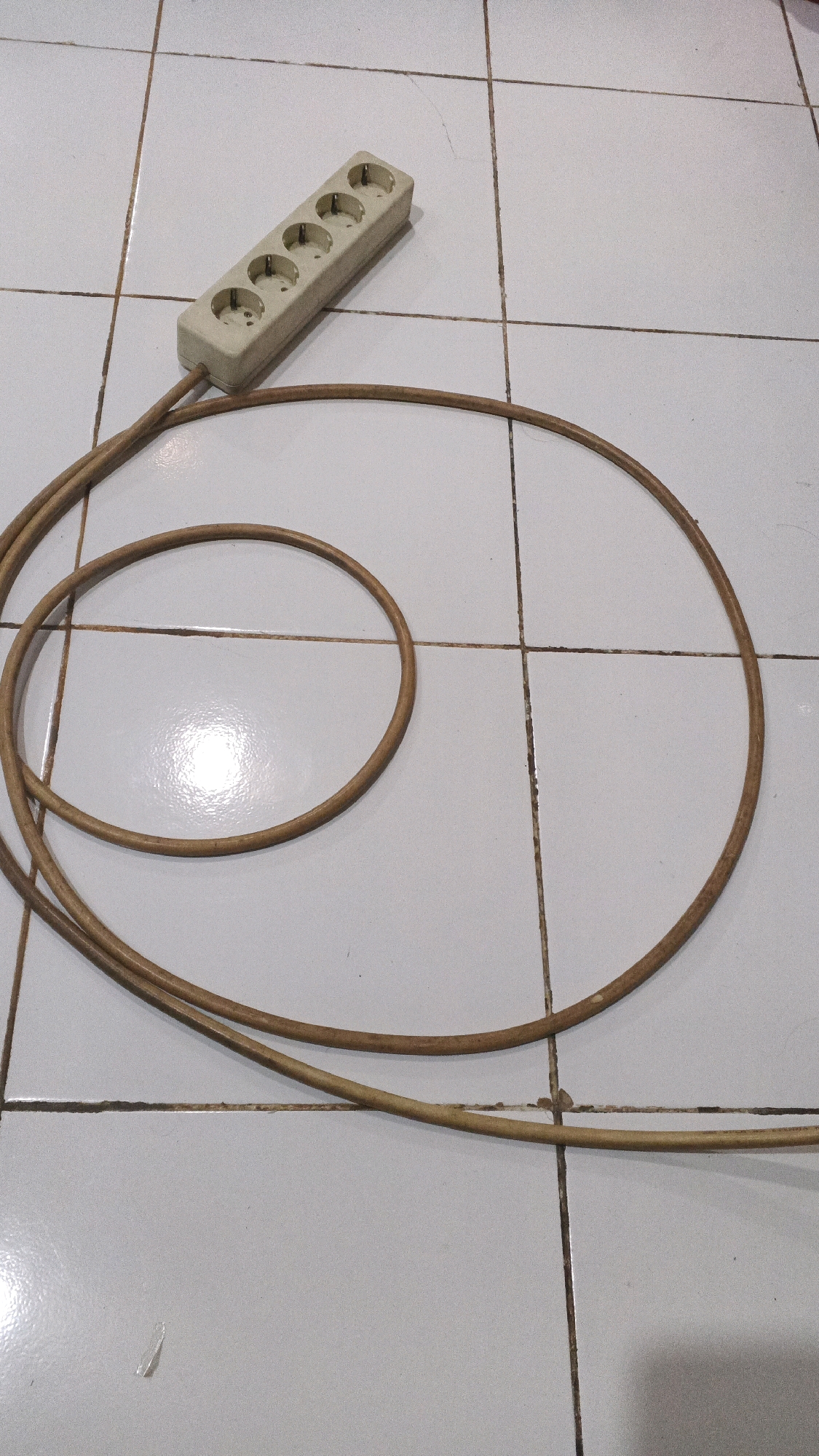 A beige multi-socket power strip with five outlets is lying on a light gray tiled floor. A brown electrical cord extends from the power strip and forms a large loop. The power strip is positioned on the upper left side of the image, angled towards the center. The cord begins at the bottom left of the power strip and curves down and to the right, forming a large circular loop before extending out of frame to the bottom right. The tiled floor has visible grout lines and some subtle dirt or scuff marks. There are no people or animals present. The lighting appears to be artificial, casting a slight shadow. This scene is located in Winong, Indonesia.
