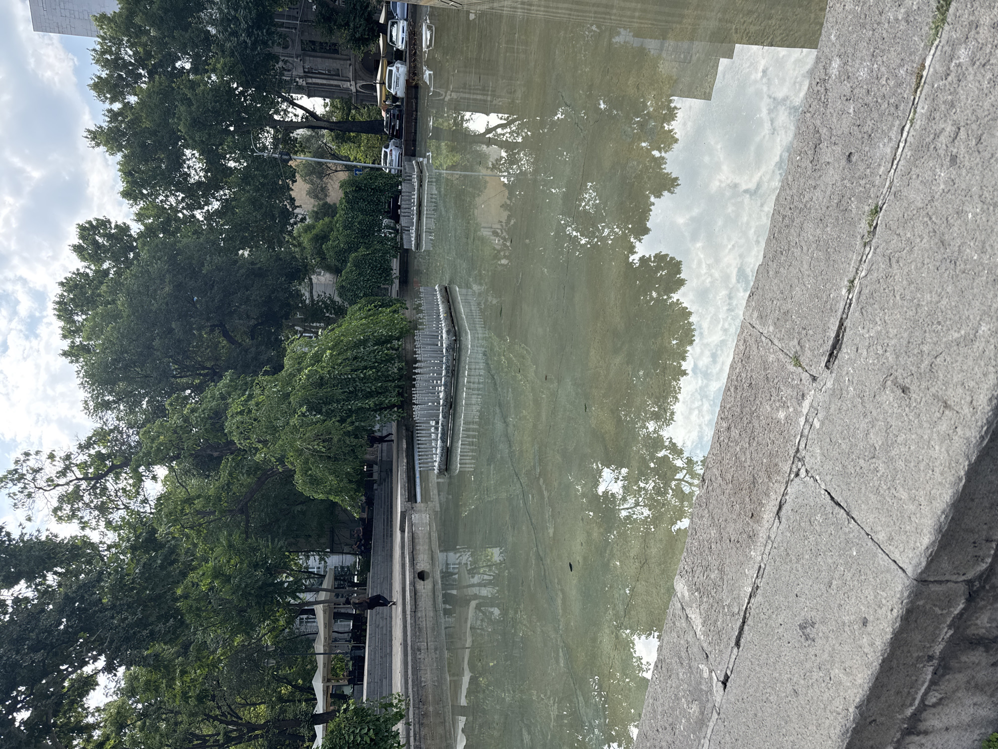 This is a medium shot, eye-level, outdoor, daytime, vertical, full, documentary photograph of a mostly empty fountain in Yerevan, Armenia. The fountain is a large, rectangular body of murky, shallow water with a light green hue. The bottom of the fountain is visible through the water, showing cracks and a dark, irregular shape that might be a leaf or debris. Two rectangular fountain heads, made of grey metal with numerous vertical prongs, are submerged in the water, one in the left half and another in the right half of the frame.
The fountain is bordered by a low, grey stone ledge with visible cracks and weathering. In the background, beyond the fountain, is a park area with lush green trees, some with weeping branches. There are also white umbrellas of an outdoor cafe and a few patrons sitting at tables. Further back, a street with parked cars, including white, black, and yellow vehicles, is visible. A tall, grey, modern building stands to the right, partially obscured by trees and a more ornate, older stone building. The sky is partly cloudy with white clouds visible reflecting in the water. Two individuals are walking in the background; one is in the mid-left area near the cafe, and another is further to the right. The overall scene depicts a quiet, daytime moment in an urban park setting.
