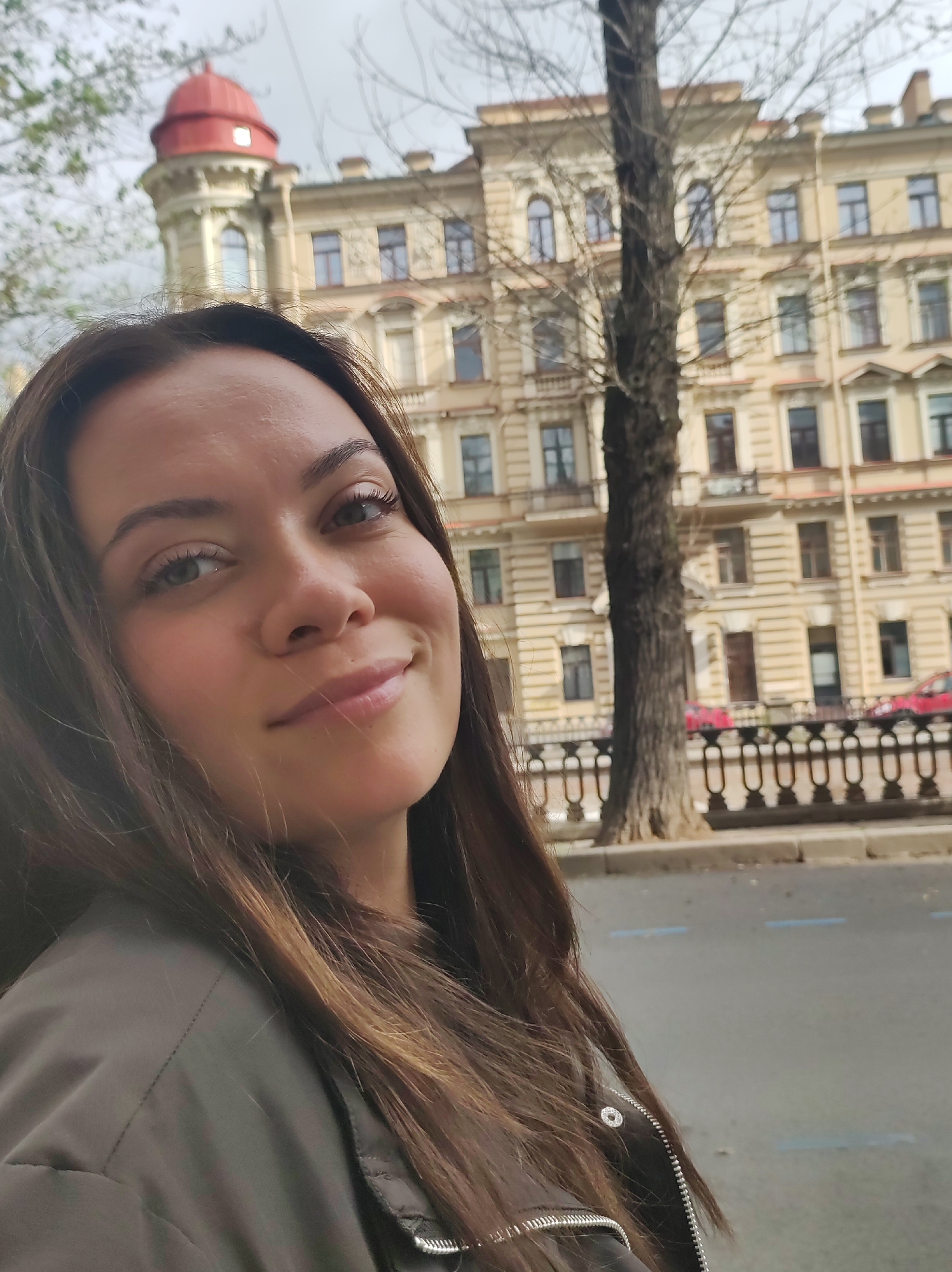 A woman with long brown hair is taking a selfie outdoors in Saint Petersburg, Russia. She is wearing a dark jacket. Behind her, a light-colored building with an ornate facade and a red dome is visible. A tree with bare branches stands in front of the building. The background also shows a street with a sidewalk and a body of water, possibly a canal or river. The sky is overcast.