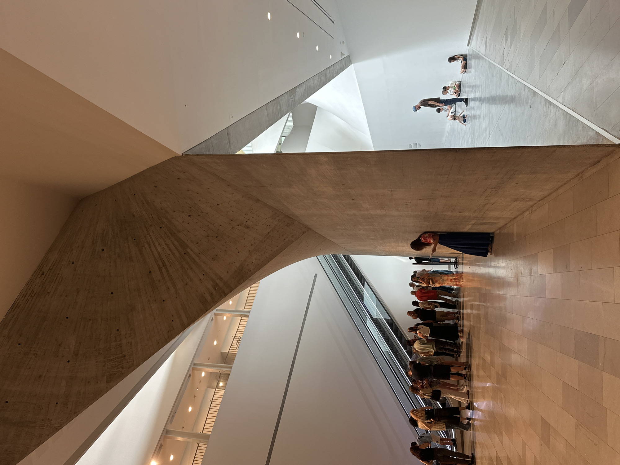 The image depicts an interior shot of a modern architectural space in Tel-Aviv, Israel. A large group of people, likely museum visitors, are lined up next to an escalator. An individual stands away from the group, apparently observing. The architectural features include exposed concrete, white walls, an angled ceiling, and a multi-story staircase visible in the background. The floor is tiled.