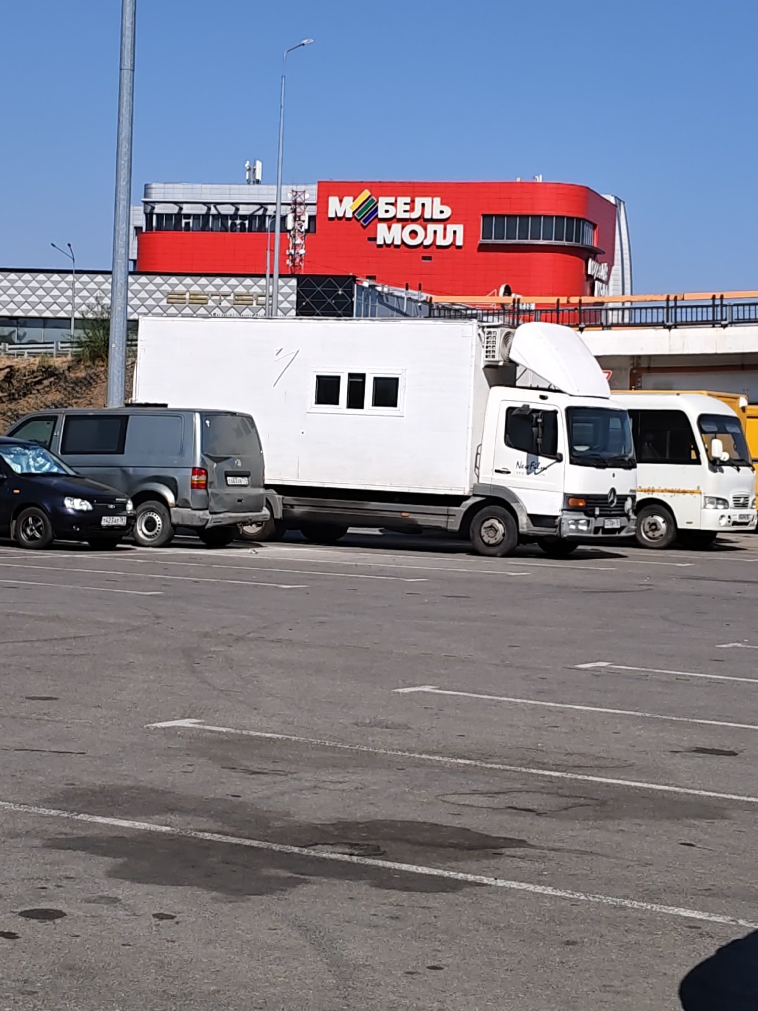 In Rostov-on-Don, Russia, a sunny day unfolds in a parking lot outside the "Mebel' Moll" shopping center, easily identifiable by its prominent red facade and Cyrillic signage. A variety of vehicles are parked, including a dark sedan, a gray van, a white truck with a cargo box, and a white minibus. The ground of the parking lot is asphalt with visible white parking lines and several dark stains, possibly from oil or water. In the background, the "Mebel' Moll" building stands out, characterized by its rounded red upper section and geometric patterns on the lower part. Several metallic poles and antennas are also visible against the clear blue sky.