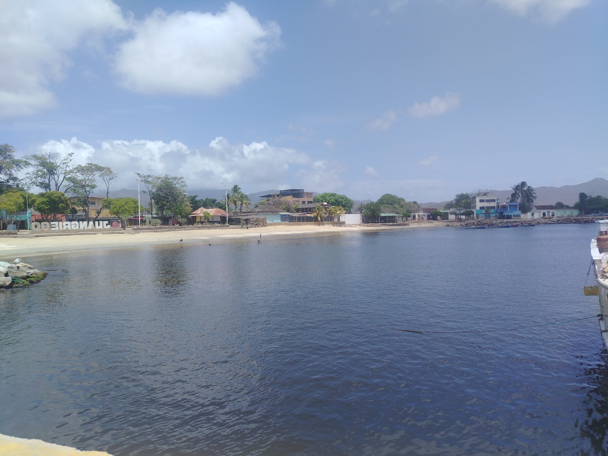 The image shows a view of the bay in Juan Griego, Venezuela, on a sunny day with scattered clouds. The water is a dark blue, with some ripples visible. In the foreground, the edge of a yellow and white structure is visible on the left and right sides of the frame. A rocky, moss-covered embankment is on the far left. A sandy beach extends across the middle ground, lined with palm trees and various buildings, including houses, a resort with a dark-tiled roof, and what appears to be a small restaurant or bar with a thatched roof. A white sign with large, red letters that spell "JUANGRIEGO" is partially visible on the left side of the beach. Several small boats are moored in the water closer to the shore on the right side of the frame, and one person is walking on the beach. In the distance, green hills rise against the horizon. The sky is a clear blue with fluffy white clouds.