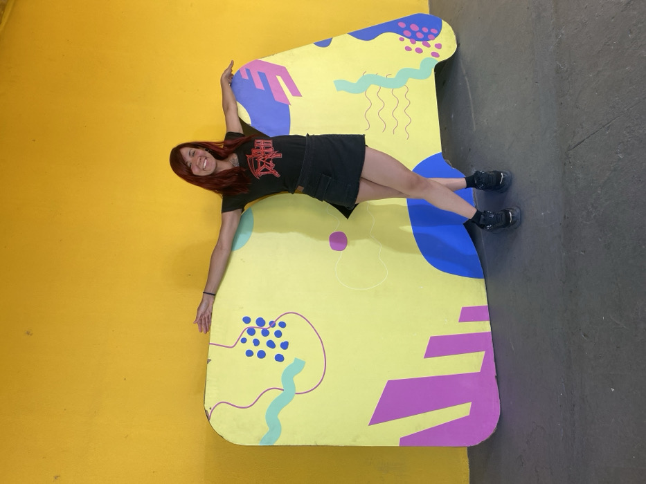 A woman with reddish hair is positioned at an angle on a large, yellow, geometric figure with blue and magenta designs. She is wearing a black t-shirt with Japanese lettering and a black skirt. Her arms are outstretched, with one hand touching a bright yellow wall and the other extended towards a gray floor. Her legs are slightly bent, with her feet planted on the gray floor. The backdrop includes the yellow wall and the gray floor, which create a visually disorienting effect.
