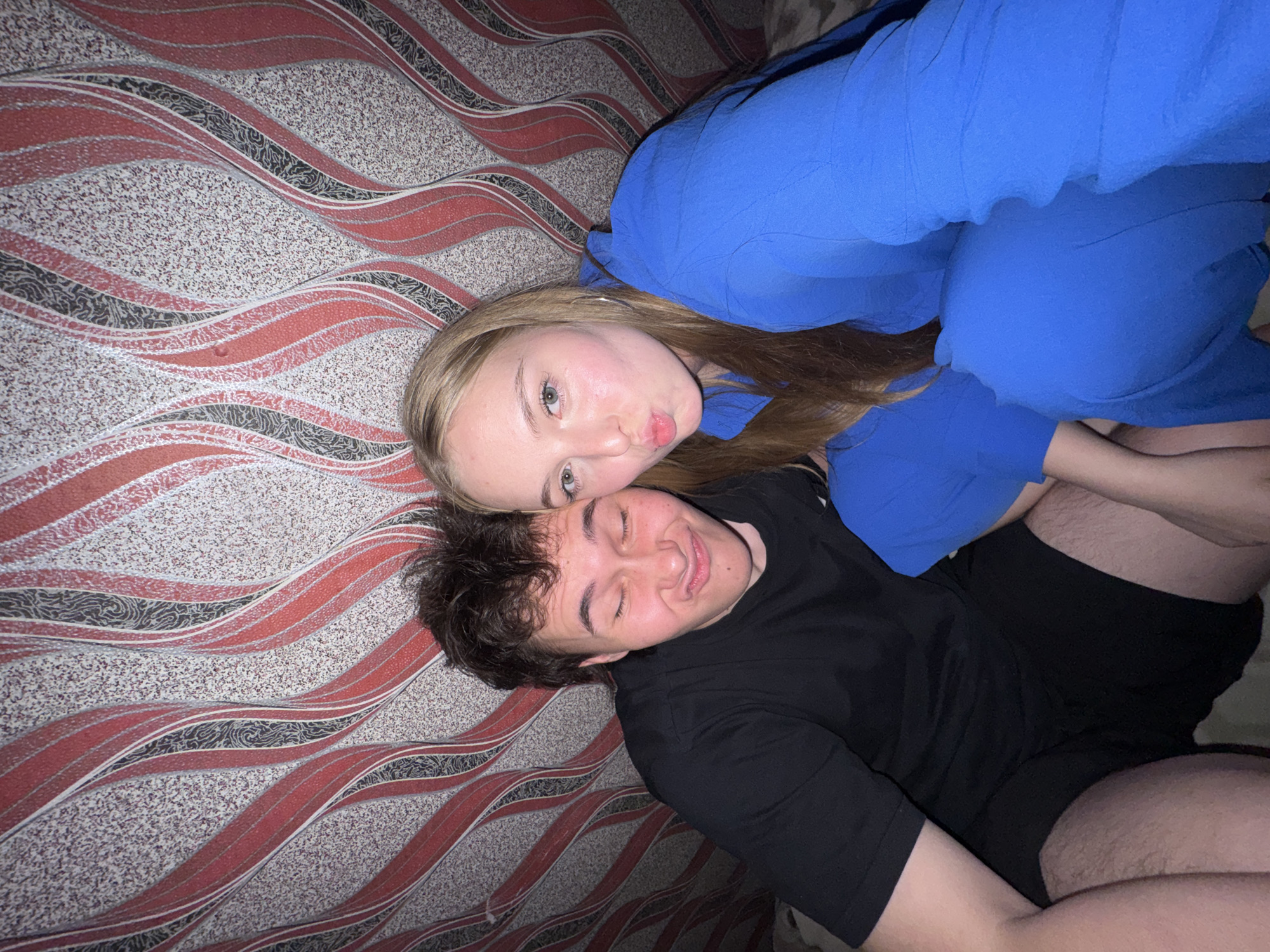 In this close-up, indoor selfie, two young individuals are framed against a vibrant, patterned wallpaper featuring wavy lines of red, black, and white. The person on the left, a young male with dark, tousled hair, squints his eyes and puckers his lips, wearing a black t-shirt and black shorts. To his right, a young female with blonde hair, also puckering her lips, looks towards the camera. She is dressed in a bright blue outfit, possibly a jumpsuit or matching top and pants. Their faces are close together, suggesting an intimate or playful moment. The background wallpaper, with its dynamic pattern and contrasting colors, fills the entire frame behind them. The lighting is warm and appears to be artificial, casting soft shadows on their faces. There is no indication of the specific location within the city of Perm, Russia.