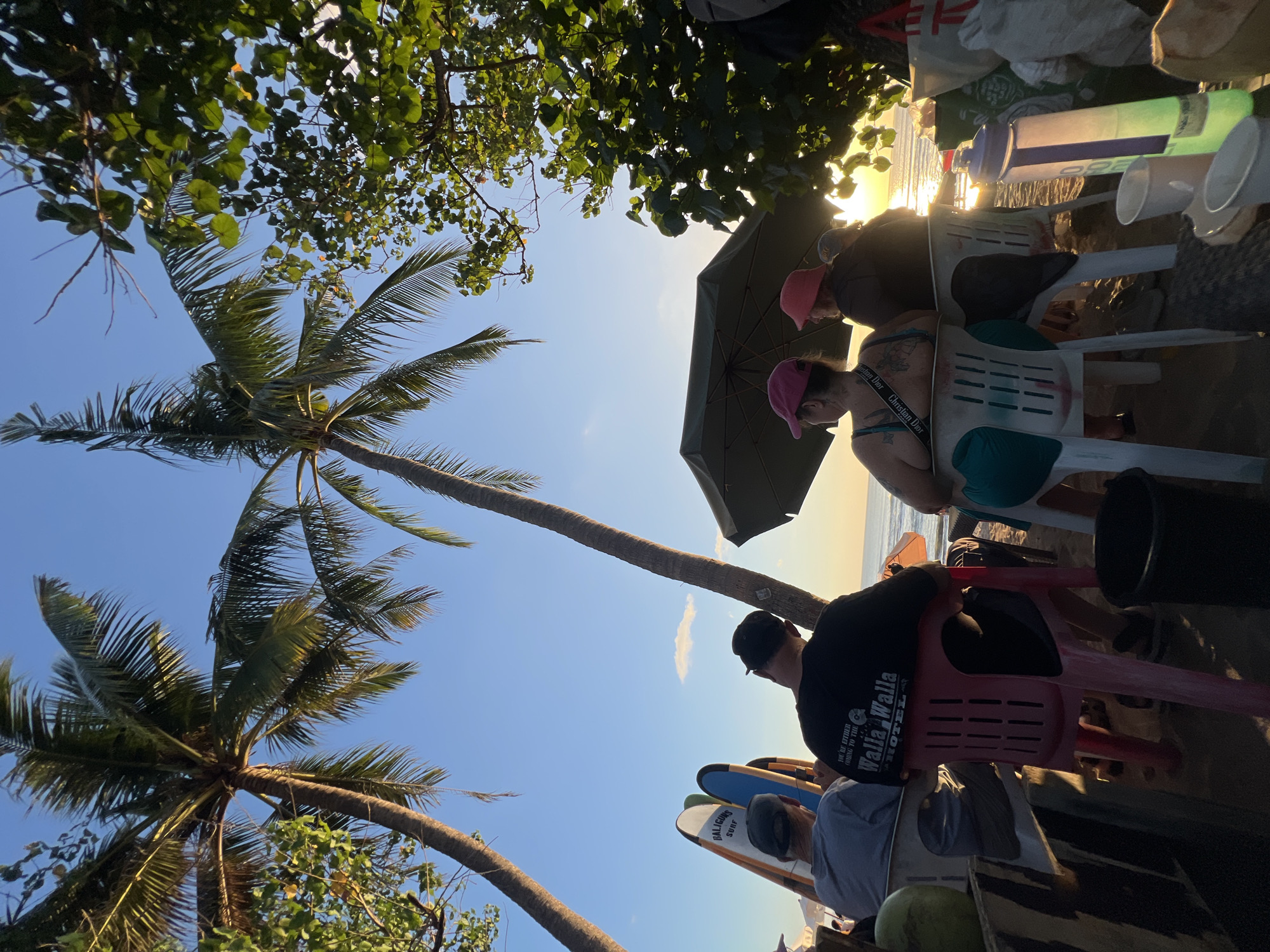 Four individuals are seated on plastic chairs on a beach in Kuta, Indonesia, facing the ocean at sunset. Two individuals are positioned in the foreground, with the person on the left wearing a grey cap and t-shirt, and the person on the right wearing a black t-shirt with text. Two other individuals are seated behind them, one wearing a pink hat and the other a red hat. A large green umbrella is set up behind them, providing shade. Two surfboards stand upright to the left of the seated individuals. Palm trees are visible in the background, framing the sunset over the ocean. The ground is sandy, with various items like bags, coconuts, and cups scattered around. The sky is clear with a few wispy clouds.