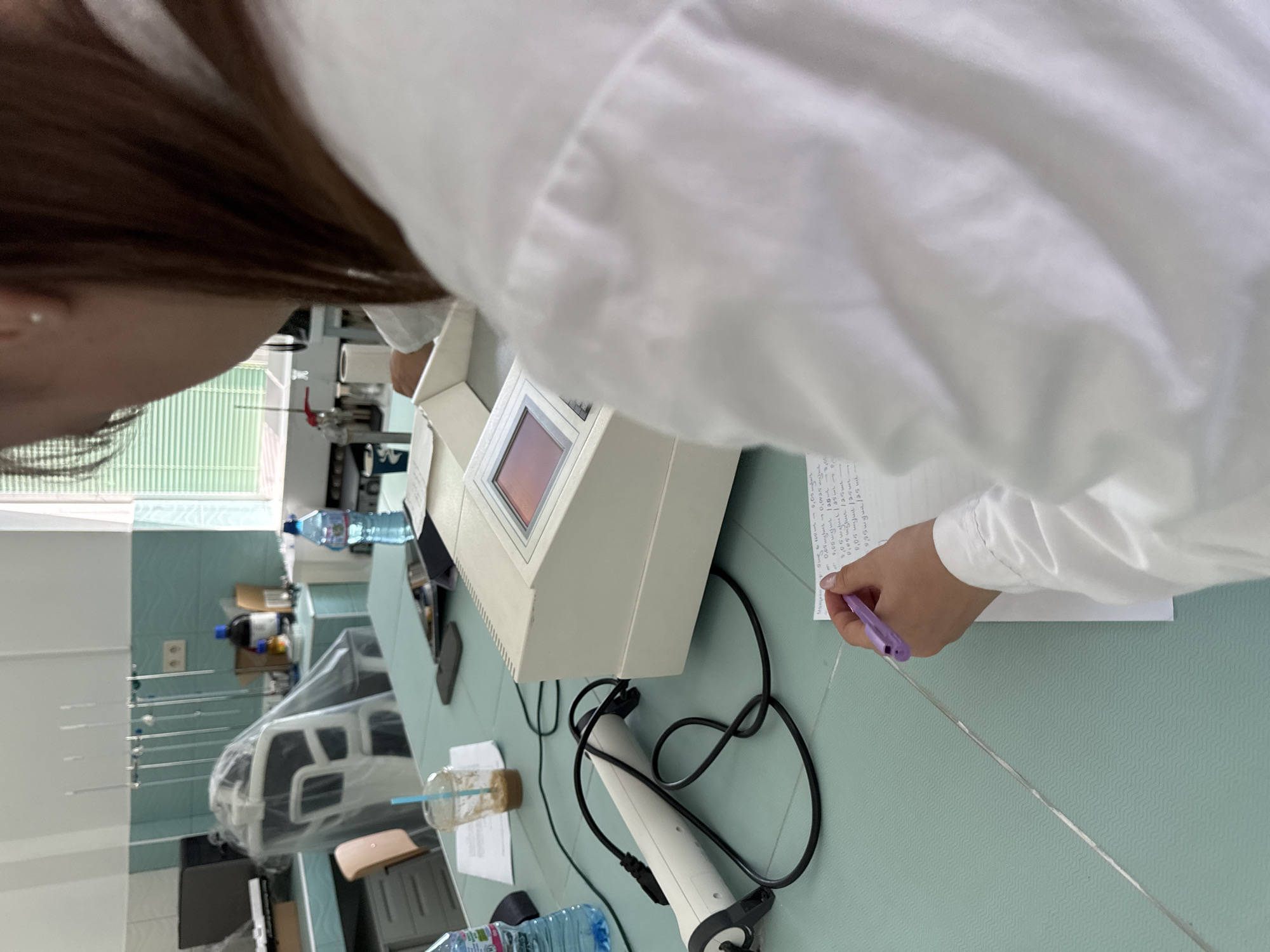 A person in a white lab coat is recording data, likely from an experiment conducted with the spectrophotometer on the table. The lab setting is indicated by the lab equipment in the background, the presence of a water bottle, and the style of the table. The image does not contain enough information to locate this lab specifically in Sofia, Bulgaria.