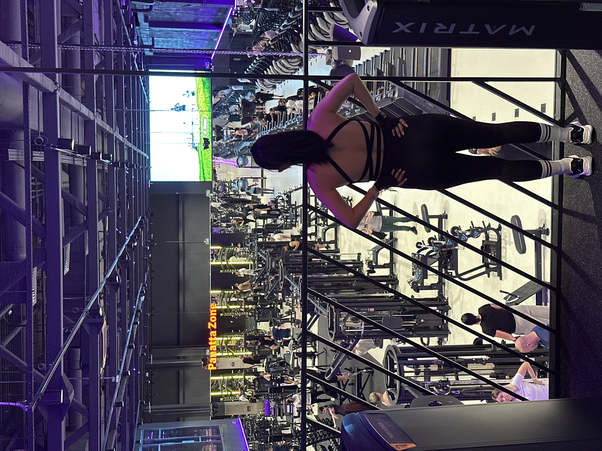 The image depicts the interior of a gym, likely in Murino, Russia. The gym appears modern with high ceilings and exposed infrastructure. Numerous people are engaged in various fitness activities using treadmills, weight machines, and other equipment. A woman in black workout attire and white-striped socks is standing on an elevated platform, seemingly stretching or adjusting her posture. A large screen displays a video of an outdoor scene, possibly a running track, further simulating an outdoor workout environment. The gym appears to be well-lit with a combination of natural and artificial lighting. The words "Panatta Zone" are visibly illuminated on a wall sign.