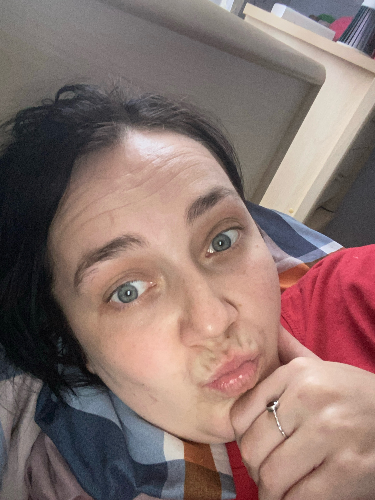 A selfie taken by a person with dark hair and light blue eyes. The person is wearing a red shirt and a ring on their right hand. The person is lying down, with a blanket and pillow visible. The background appears to be an indoor setting with a wooden shelf and some books. The lighting is soft and appears to be natural light coming from a window.