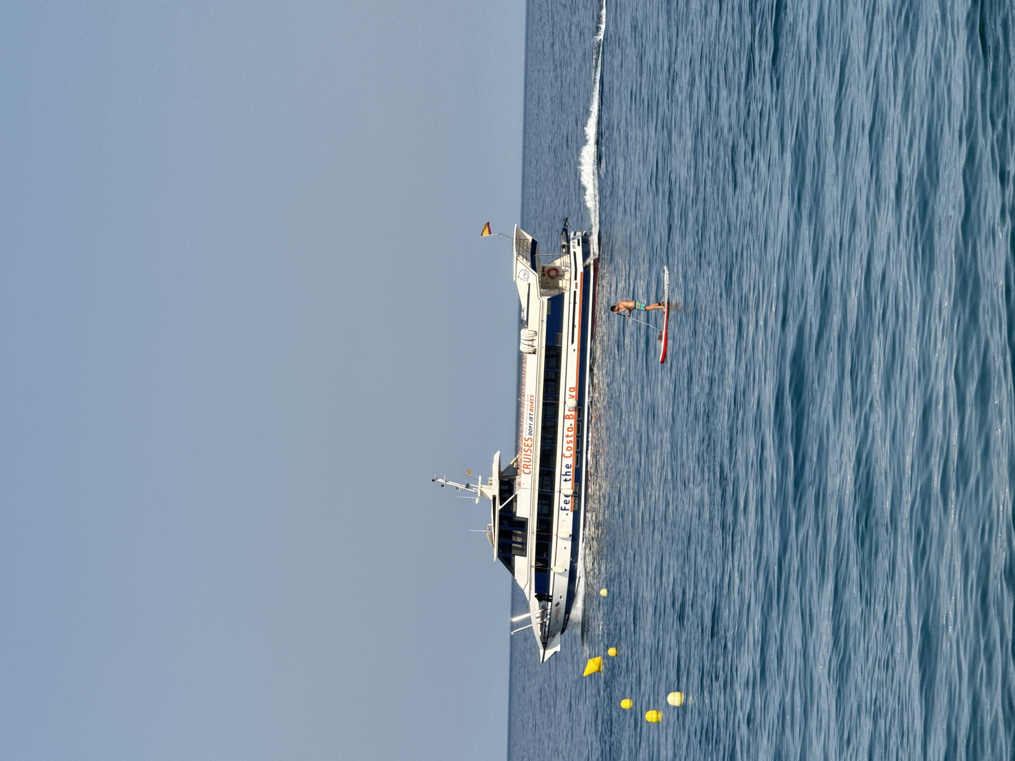 A person is paddleboarding on a body of water. The person is standing on a red paddleboard and is holding a paddle. The person is wearing blue and green swim trunks and is facing away from the camera. Behind the paddleboarder, a white boat with blue trim is moving through the water. The boat has text on its side that says "CRUISES DOFI JET BOATS" and "Fee the Costa-Bra". The boat has a Spanish flag at the stern. In the water to the left of the paddleboarder, there are several yellow buoys. The scene appears to be in the sea, possibly near Blanes, Spain, given the text on the boat. The sky is clear and blue.