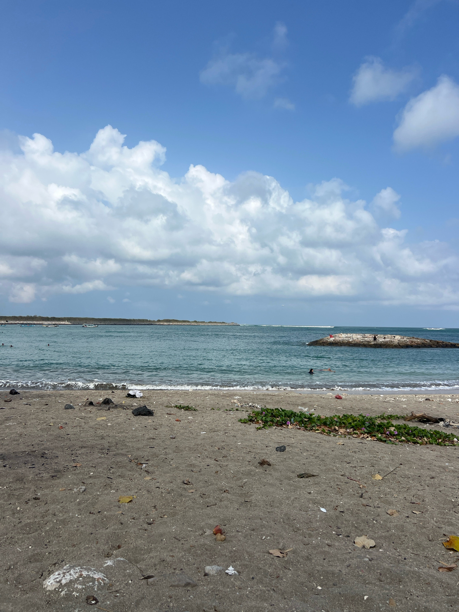 This is a photograph of Kuta Beach in Bali, Indonesia. The image captures a wide view of the shoreline under a bright blue sky dotted with fluffy white clouds. The sand in the foreground is a dark gray and appears somewhat cluttered with debris, including rocks, leaves, and some white plastic.
The ocean is a tranquil turquoise color, with gentle waves lapping at the shore. In the middle distance, a man-made breakwater, constructed from large stones, juts out into the water. Several people are visible near or on the breakwater, with some appearing to be fishing or relaxing. A few individuals are also in the water, seemingly swimming or wading.
Further out, a line of boats is visible along the horizon, suggesting a coastal village or harbor. The overall scene conveys a relaxed, natural atmosphere typical of a beach destination.