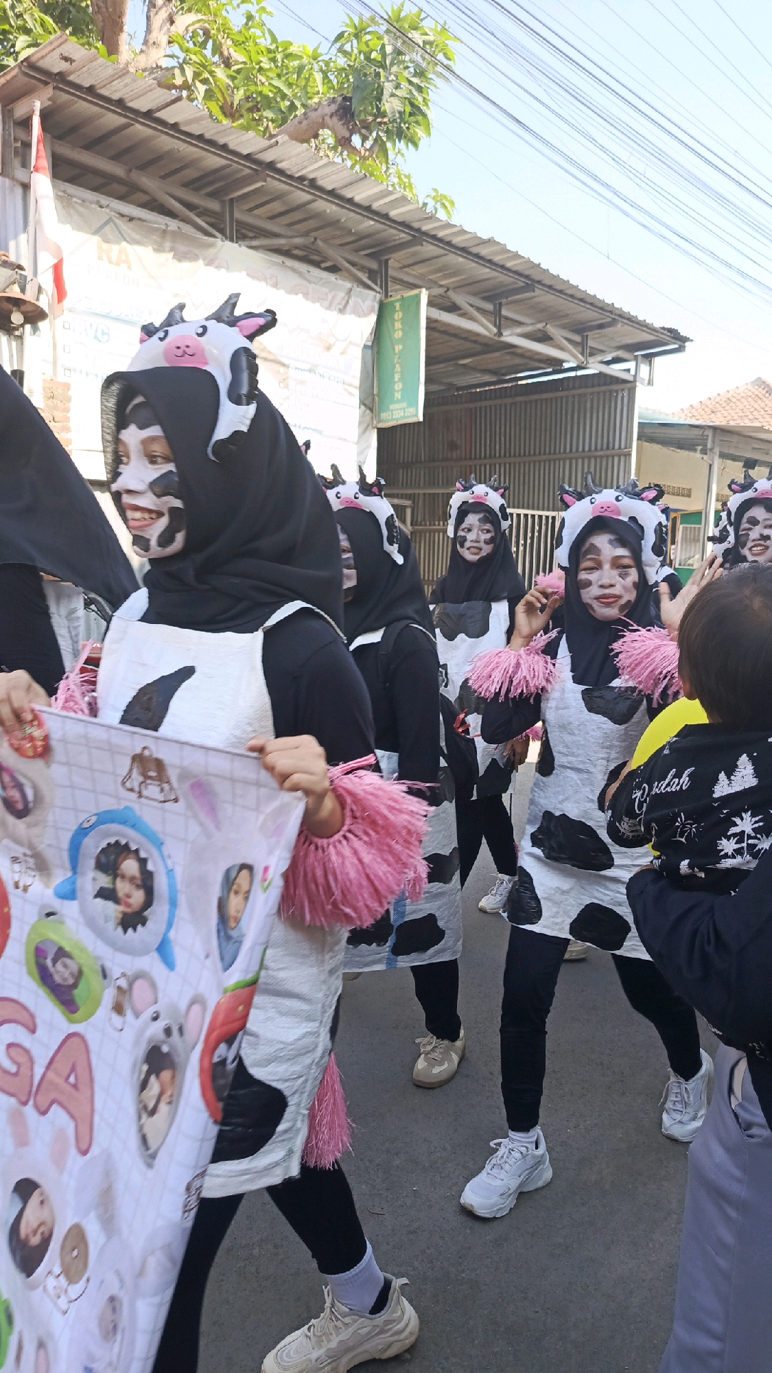 In Pekauman Kulon, Indonesia, a group of individuals, possibly children or young adults, participate in a parade or performance. They are dressed in cow-themed costumes, featuring black and white spotted outfits, cow-ear headbands, and face paint designed to resemble cow markings. Several individuals are holding a banner adorned with images, including faces and cartoon characters. The scene takes place outdoors on a street, with buildings and power lines visible in the background, suggesting a village or town setting. The sky is clear and bright, indicating daylight.