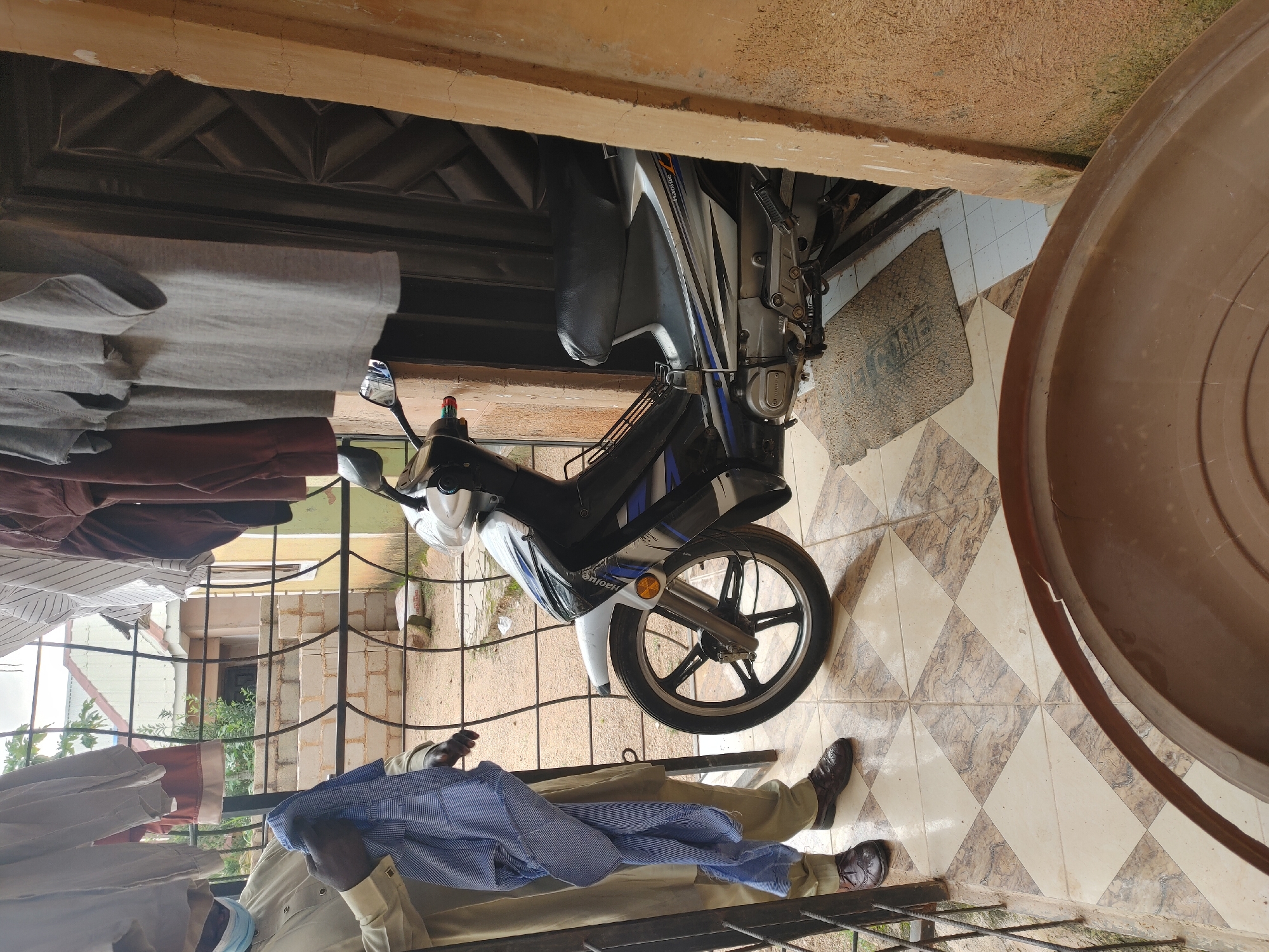 A person wearing a face mask and a light-colored robe is holding a blue and white checkered shirt. The person is standing on a tiled patio next to a parked motorcycle. The motorcycle is black and silver with blue accents. A dark brown textured door is visible behind the motorcycle. Several garments are hanging on a clothesline in the background. The scene appears to be set in an outdoor area with some construction visible.