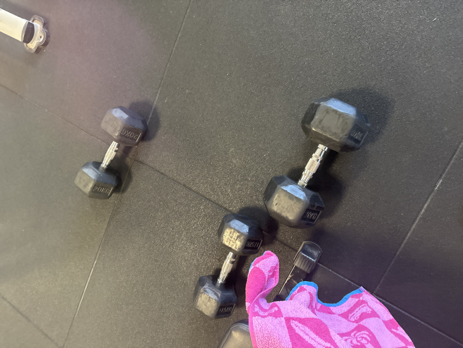 The scene is located in a gym in Neuville-aux-Bois, France, on a dark, textured rubber floor composed of interlocking mats. Three hexagonal dumbbells are present: one 20KG dumbbell positioned in the upper right, a 17.5KG dumbbell in the mid-left, and a 9.0KG dumbbell in the lower right. A pink and blue patterned towel lies partially visible in the lower-left quadrant, covering part of a black spray bottle with its nozzle pointed left. A segment of a metal base from gym equipment is visible in the upper right background. No individuals are present, and no actions or interactions beyond static placement are depicted.