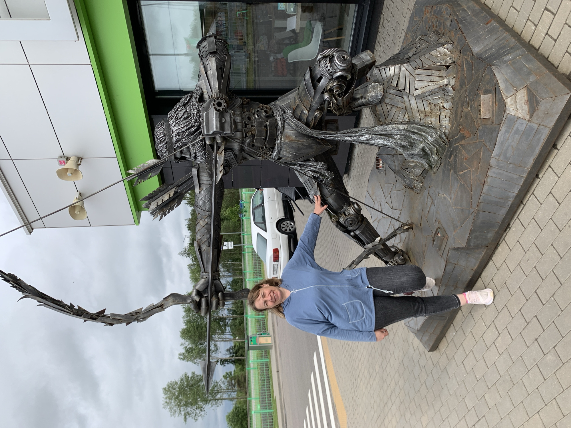A woman stands next to a large metal sculpture of an archer in the city of Дзешчанскі сельскі Савет, Belarus. The sculpture, made of various metal pieces, depicts an archer in mid-shot, with a bow drawn and an arrow nocked. The archer has a detailed face and body, with armor-like plating and a cape made of metal. The sculpture is mounted on a stone base. The woman, wearing a blue hooded jacket and dark jeans, smiles at the camera and rests her hand on the sculpture. In the background, a white car is parked, and a building with a green trim is visible. The scene is outdoors, with a paved walkway and a cloudy sky.