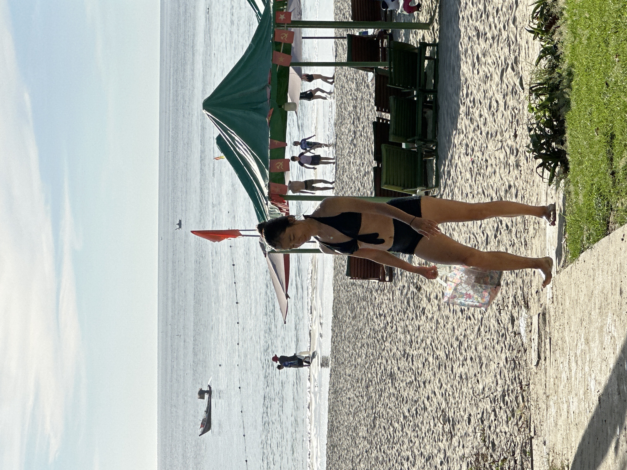The image depicts a beach scene in Da Nang, Vietnam, under a bright, sunny sky. In the foreground, a woman in a black bikini walks along a sandy path. She is carrying a transparent tote bag with a floral pattern. The beach is covered in sand with some patches of green vegetation at the bottom right. In the middle ground, several green beach umbrellas with red flags are set up. Behind the umbrellas, a group of people, including children, are wading in the shallow ocean water. Further out in the sea, a small fishing boat is visible. The ocean stretches to the horizon, meeting a pale blue sky with wispy clouds. The overall atmosphere is one of a relaxed beach day.