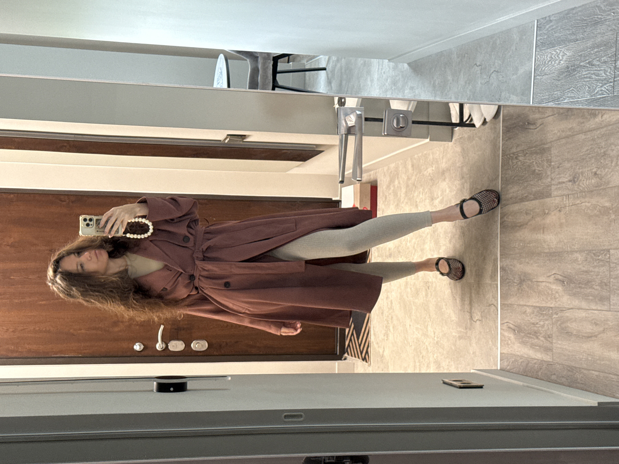A young woman is captured in a mirror selfie within an indoor setting, likely a hallway or entryway in Moscow, Russia. She has long, wavy dark brown hair and is looking directly towards the camera reflected in her smartphone. She is wearing a long, reddish-brown or plum-colored trench coat, belted at the waist, over light grey leggings and black flat sandals with a woven or mesh upper.
Her left hand holds a light-colored smartphone, adorned with a white beaded strap, which she is using to take the photo. The reflection shows a dark brown wooden door with a silver handle and two round lock mechanisms to her left. Below the mirror, a small black cylindrical object, possibly a smart speaker, is visible on a light-colored surface. The floor features a clean design with a combination of light brown and grey wood-look tiles. To the right, a small portion of a black and white geometric patterned mat is visible, along with another small, rectangular object on a ledge. The overall impression is a modern, well-lit residential or hotel interior. No discernible text is visible.