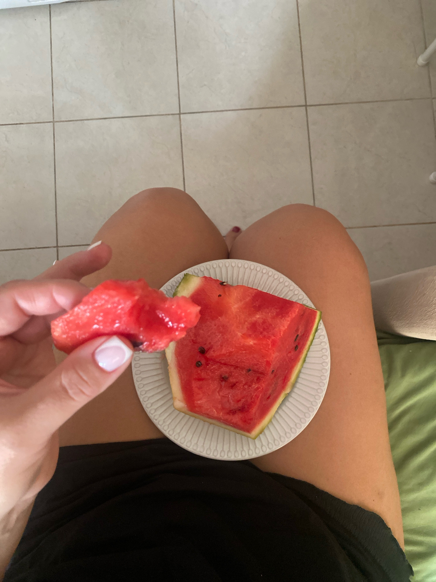 A person is sitting on a bed with a plate of watermelon. The person is holding a piece of watermelon in their hand, about to eat it. The background is a tiled floor. The person is wearing a black top. The watermelon is a slice of watermelon. The plate is white and has a pattern around the rim. The bed is covered with a green blanket. There are no people or objects in the city of Sochi, Russia, visible in the image.