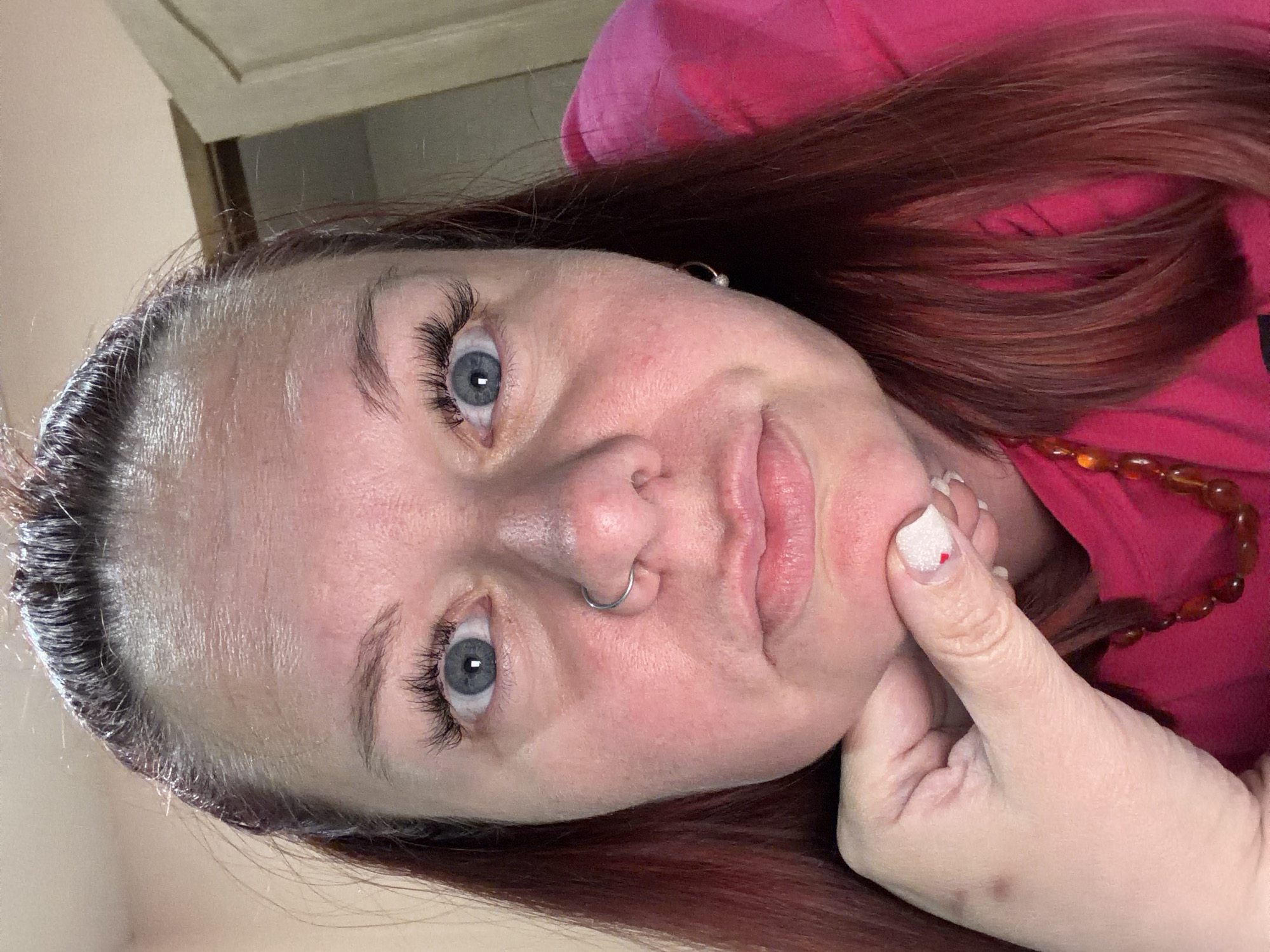 The image shows a woman with red-brown hair and blue eyes. She has fair skin and a silver nose ring. She is wearing a pink top and a necklace with amber-colored beads. Her hand is resting under her chin. The background appears to be a room with light-colored walls. There is no information available to conclude that the image was taken in Chaikovsky, Russia.