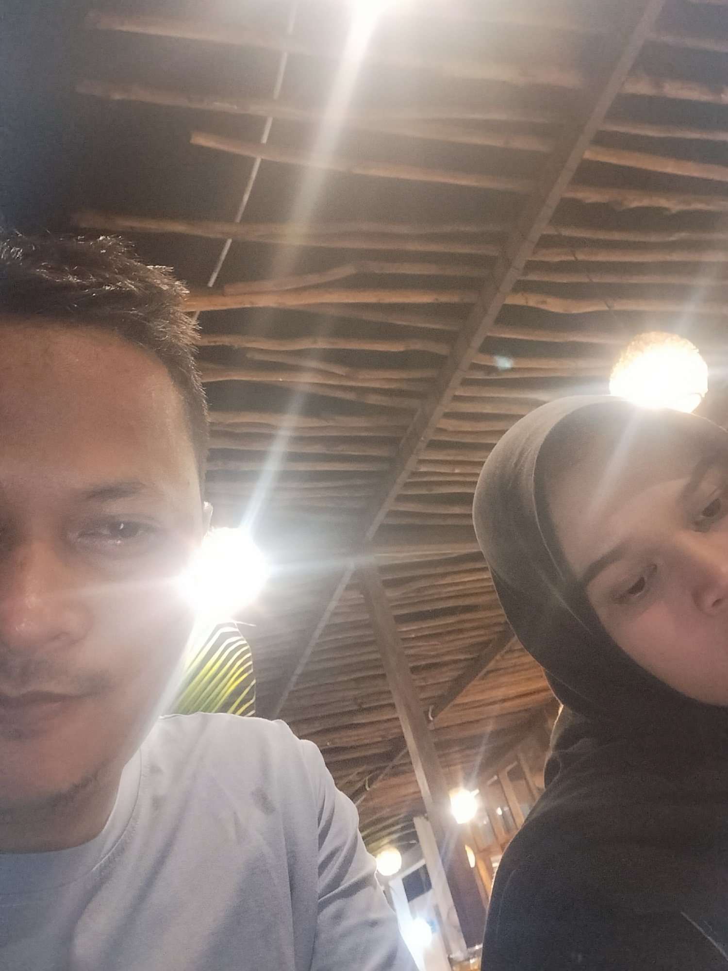 An indoor, tilted portrait features two individuals, a man and a woman, framed against a background of rustic wooden beams on a ceiling. The setting appears to be a dimly lit establishment, possibly a restaurant or cafe, in Keudekruenggeukueh, Indonesia, as suggested by the artificial lighting being the primary light source, indicating evening or nighttime.
On the left, partially visible from the forehead to just below the mouth, is a man with dark hair and what appears to be light stubble. He wears a light-colored t-shirt which shows some dampness or sweat stains on the shoulders and chest. His face is heavily backlit by a bright light source to his right, creating a significant lens flare that obscures part of his face and eye.
To the right, also partially visible, is a woman wearing a dark hijab or hooded garment. Her face is similarly affected by a bright light source directly above her, casting a strong flare. Her eyes are partially visible, looking slightly downwards or away from the camera.
The ceiling above them is constructed from numerous horizontal wooden beams or logs, creating a textured, natural aesthetic. Several bare or partially obscured light bulbs are visible in the background, emitting bright, circular glows, with one particularly prominent spherical light fixture visible near the woman's head. The overall mood is warm due to the lighting, but the subjects' expressions are not clearly discernible due to the harsh backlighting and partial views. No visible text is present in the image.
