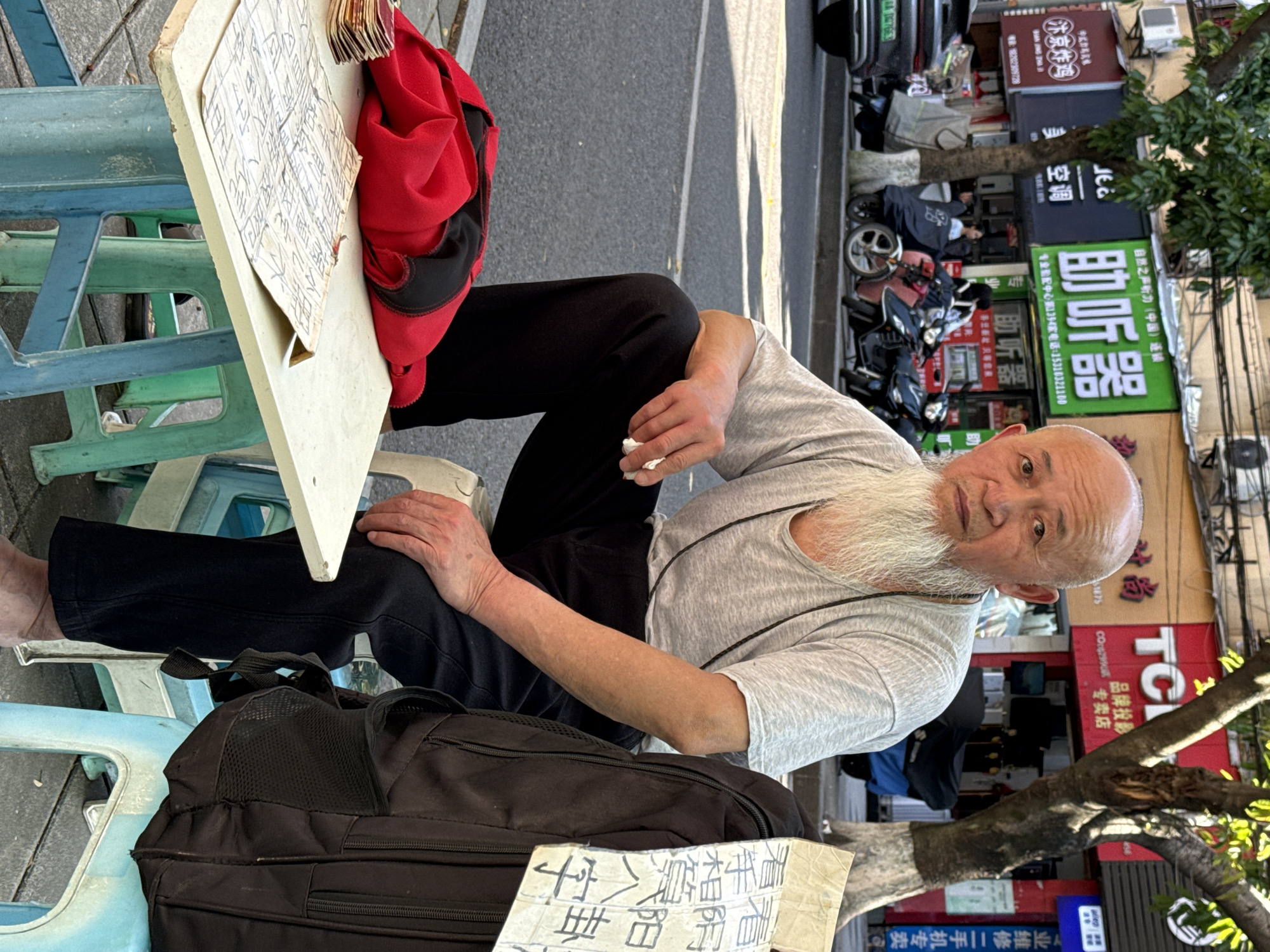 The image shows an older Asian man with a shaved head and a long, white beard sitting on a low blue plastic stool at an outdoor workstation consisting of a white plastic folding table. He is wearing a grey t-shirt and black pants. His bare foot is visible on the stool. The table has a paper with Chinese characters on it and a red bag. A black backpack is on the ground next to his stool. He is holding a tissue in his right hand and looking at the camera. The background includes a street scene with motorcycles and buildings. Storefront signs are visible with Chinese characters. A sign reading "助听器" (hearing aids) is prominent. There's also another sign that reads "汴京炸鸡" (Bianjing Fried Chicken). Based on the signs "自然之声听力(中国)连锁" (Nature's Voice Hearing (China) Chain) and other storefront signs, this appears to be in a commercial area of Beibei, China. Other signs can be read as "看手相算八字" and "看阴阳卦", suggesting this man is a fortune teller, specifically through palm reading and Chinese astrology, or maybe a geomancer of some kind. A tree is also partially visible in the background.
