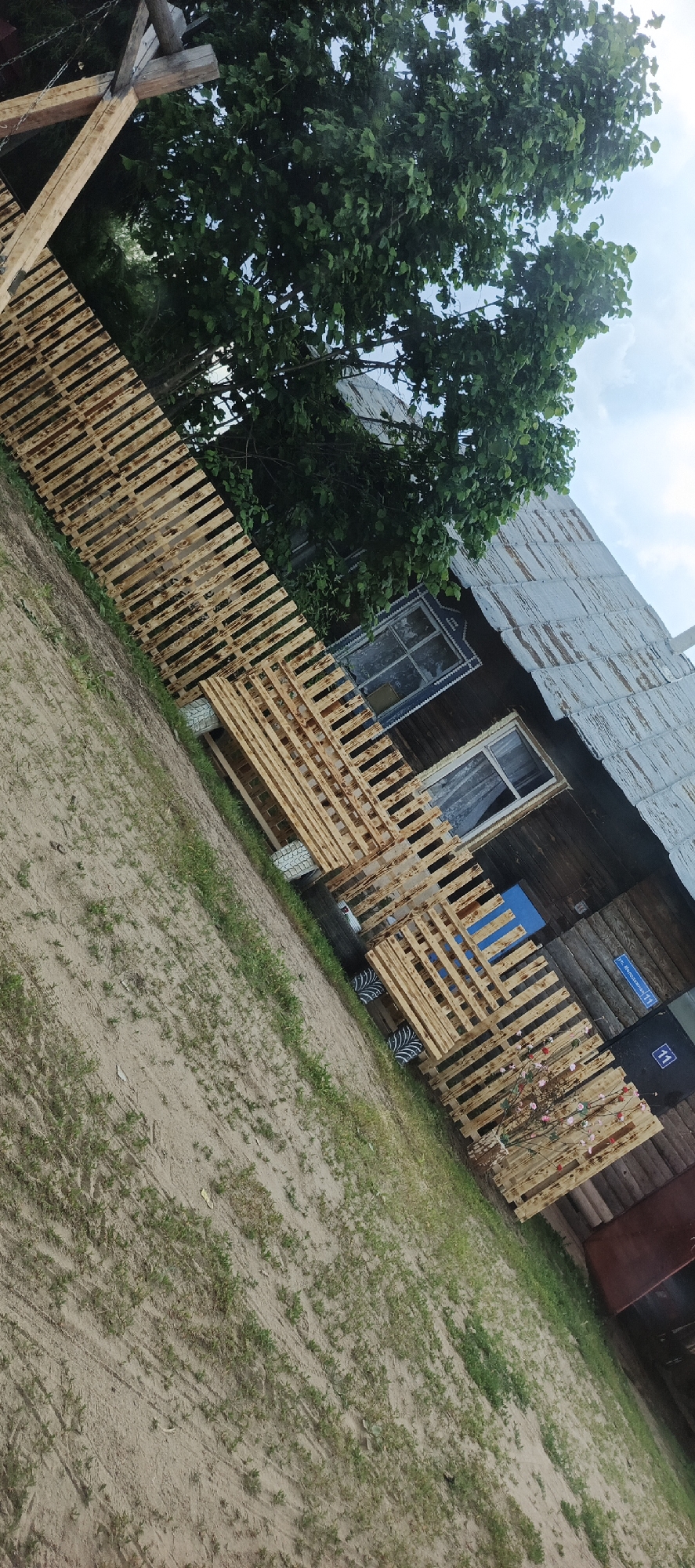 The image depicts an outdoor scene, possibly in the village of Garevaya, Russia, with a wooden house and a fence constructed from pallets. The house has a weathered, corrugated metal roof and wooden walls, with two visible windows. One window has intricate blue and white trim, while the other has sheer white curtains. The fence, made of multiple wooden pallets, runs diagonally across the foreground and appears to be a DIY project. Some of the pallets are stacked, and one section has a decorative element of branches with pink and white flowers. The ground is a mixture of sand and sparse green grass, with tire fragments visible near the base of the pallet structure. A large green tree with dense foliage overhangs the scene, with a cloudy sky visible in the background. A blue street sign with white text and numbers is attached to the house wall. There are no people visible in the image.