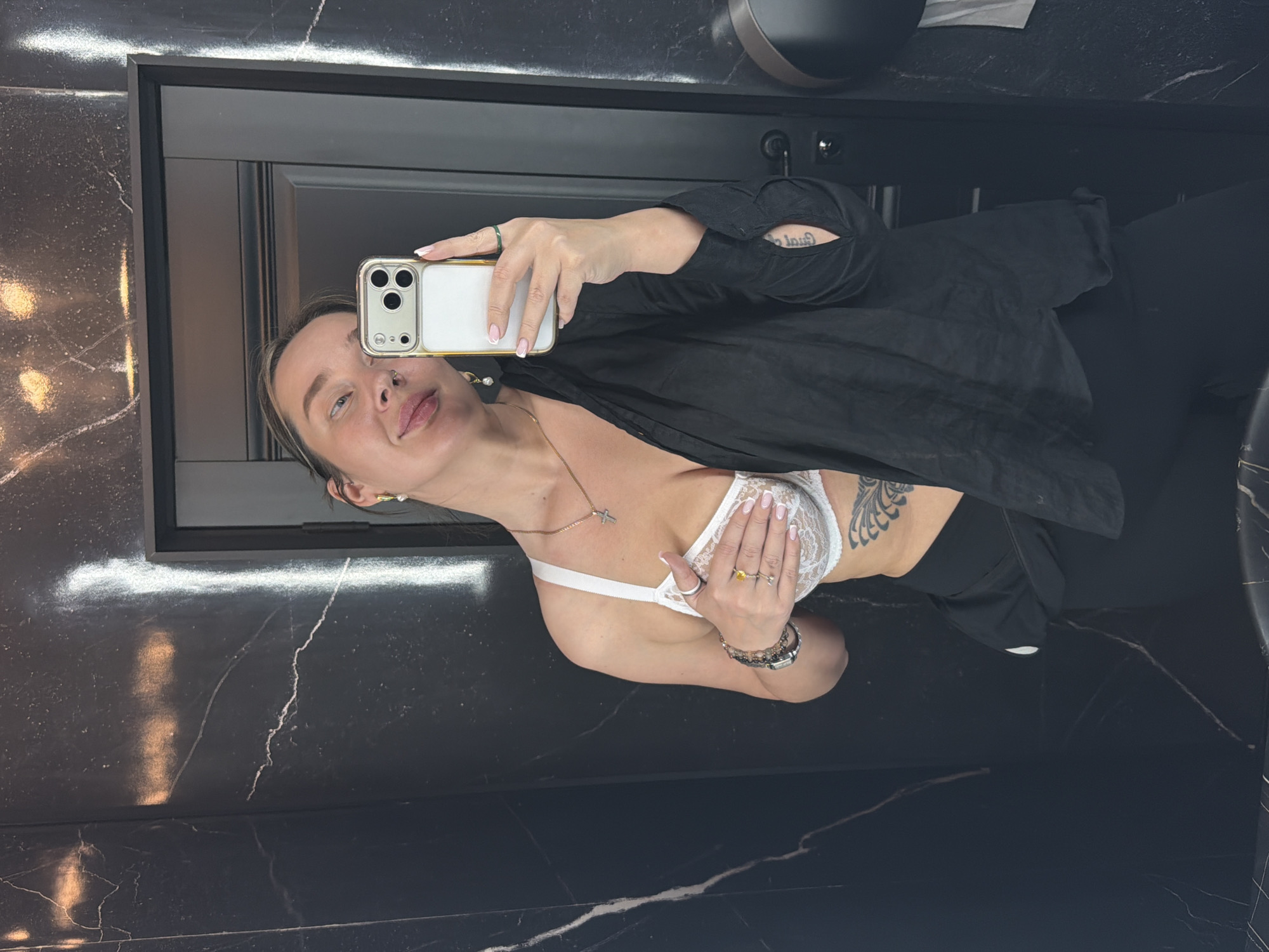 A woman is taking a selfie in a bathroom. She is wearing a white bra, black pants, and a black top. She has a tattoo on her side. The bathroom has black marble walls.