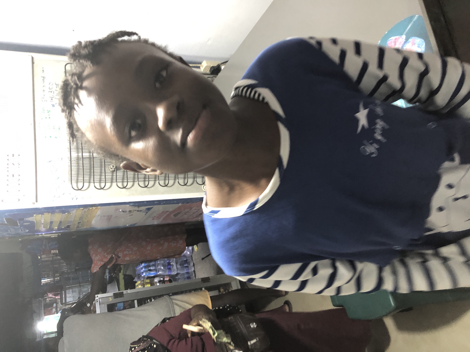 In a market stall in Ado, Nigeria, a young girl with braided hair gazes forward, her expression neutral. She wears a blue and white striped long-sleeved shirt with a graphic design on the front. Behind her, a metal refrigerator is visible, its door adorned with yellow tape. The background suggests a bustling environment, with other individuals and merchandise present, including bottles of water on shelves and a person in a red floral dress. The lighting is dim, typical of indoor market settings, casting shadows that add depth to the scene.
