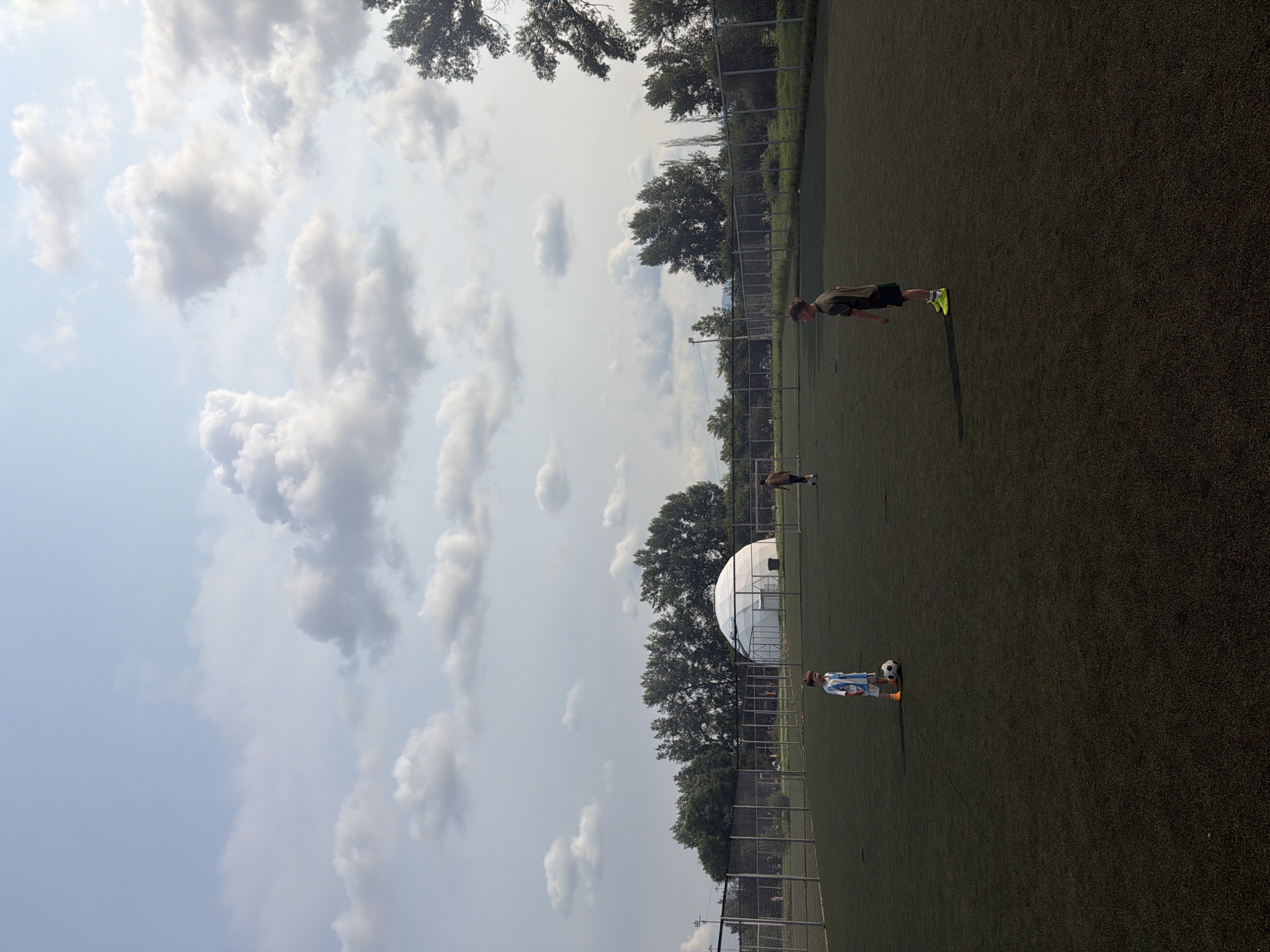 The image depicts an outdoor artificial turf sports field, likely a soccer field, in Krasnoyarsk, Russia. The field is bordered by a chain-link fence. In the background, a large, white, dome-shaped structure, possibly a sports hall or arena, is visible, with a line of trees behind it. The sky is partly cloudy with scattered white and gray cumulus clouds.
In the foreground and mid-ground, several individuals are present on the field. On the left side of the field, a young boy wearing a blue and white striped soccer jersey and shorts, along with orange cleats, stands next to a soccer ball. To the right of him, another young boy in a dark green t-shirt and black shorts, with bright yellow cleats, is positioned facing away from the camera. Further in the background, near the fence, a person wearing a brown shirt and dark pants is walking. Other indistinct figures can be seen further back near the fence and the dome structure. The lighting suggests it is daytime, possibly late afternoon or early evening, given the shadows.