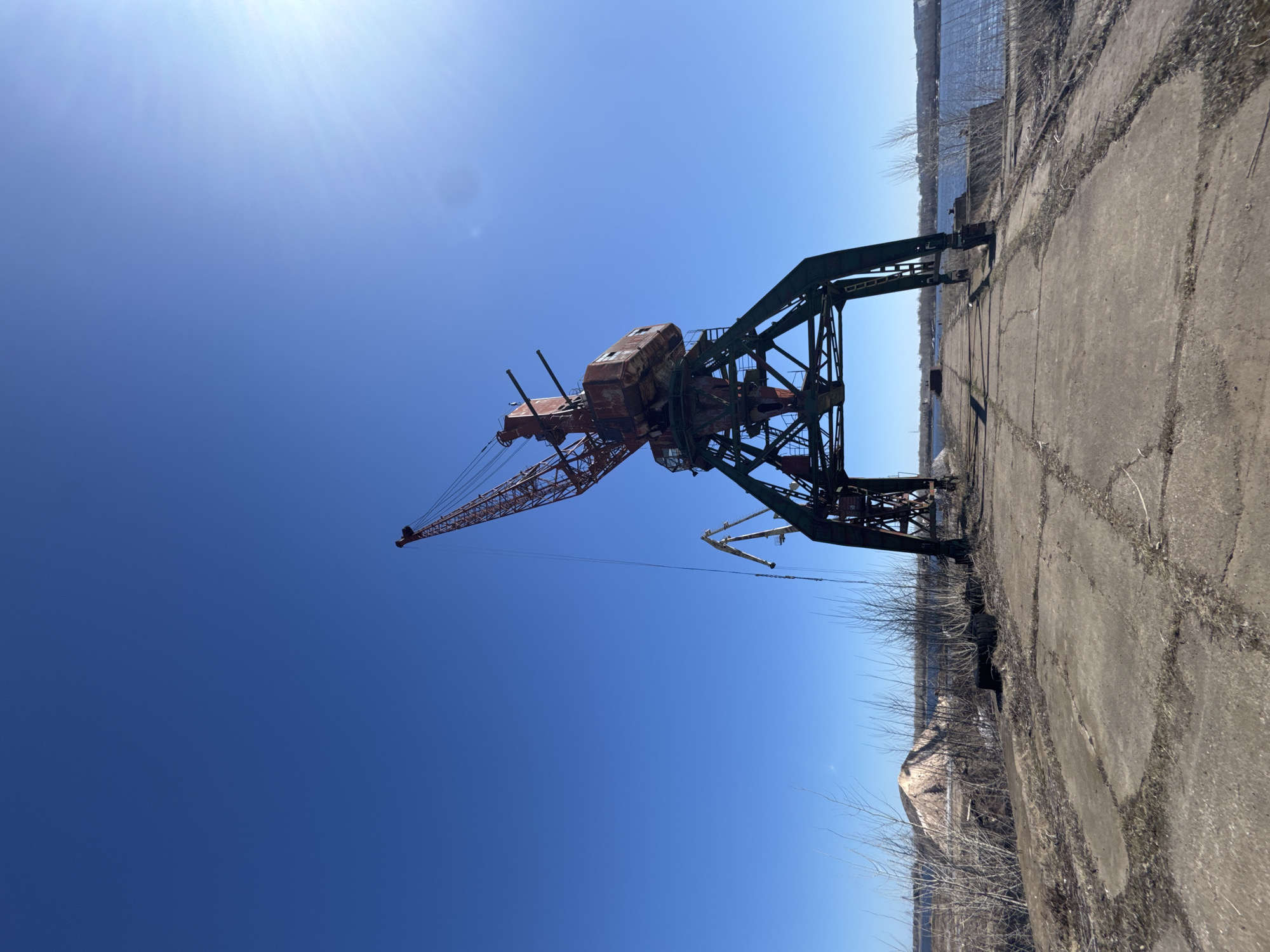 An abandoned industrial crane, located in Chaikovsky, Russia, stands on a cracked concrete platform under a clear, blue sky. The crane is predominantly rusted red with a green support structure. A body of water is visible in the background beyond the edge of the concrete platform. The platform is cracked and shows signs of disrepair.